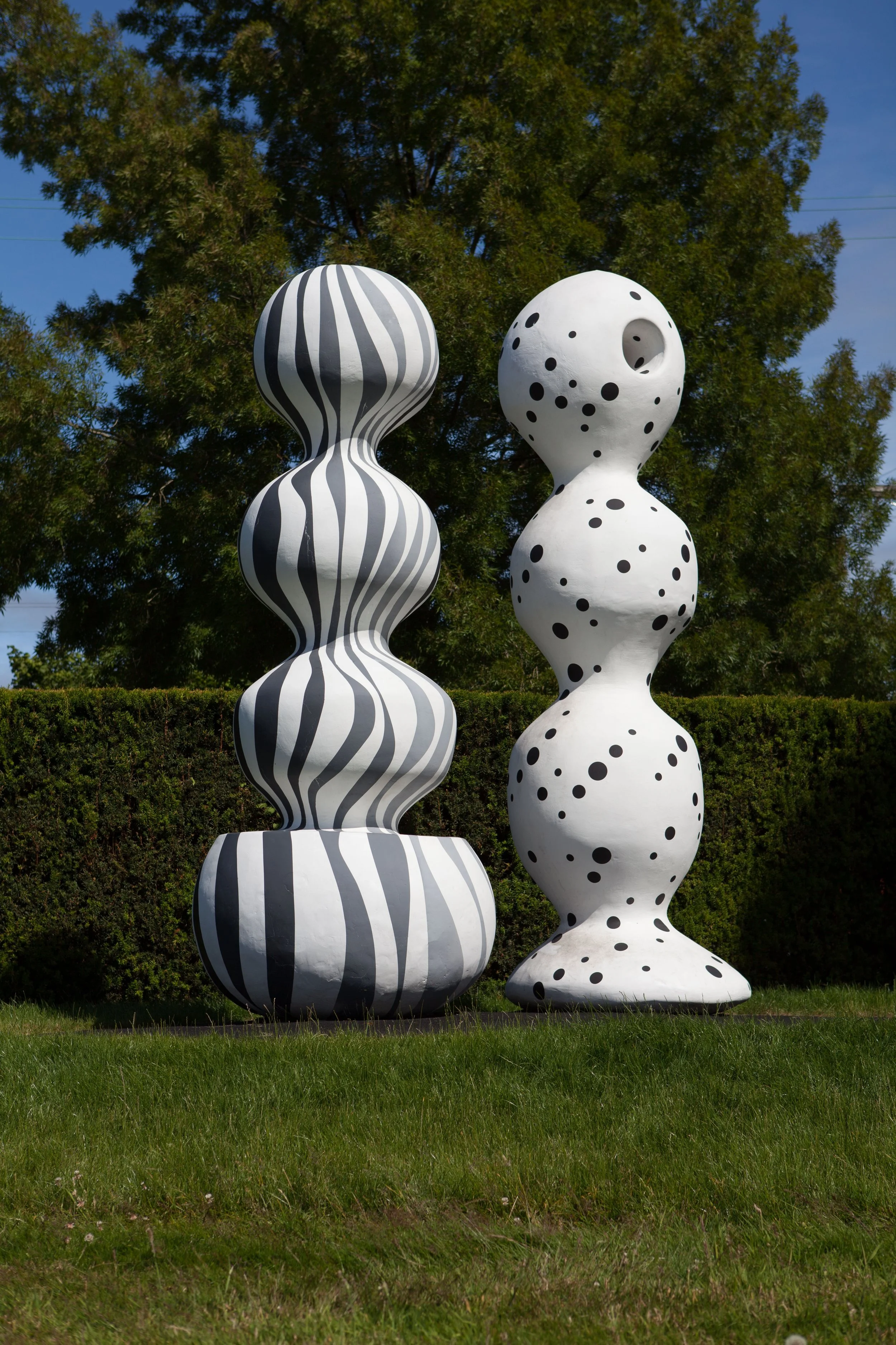 Two large modern sculptures on grass, one with black and white wavy stripes and the other with black polka dots, set against a backdrop of green trees and a bright blue sky.