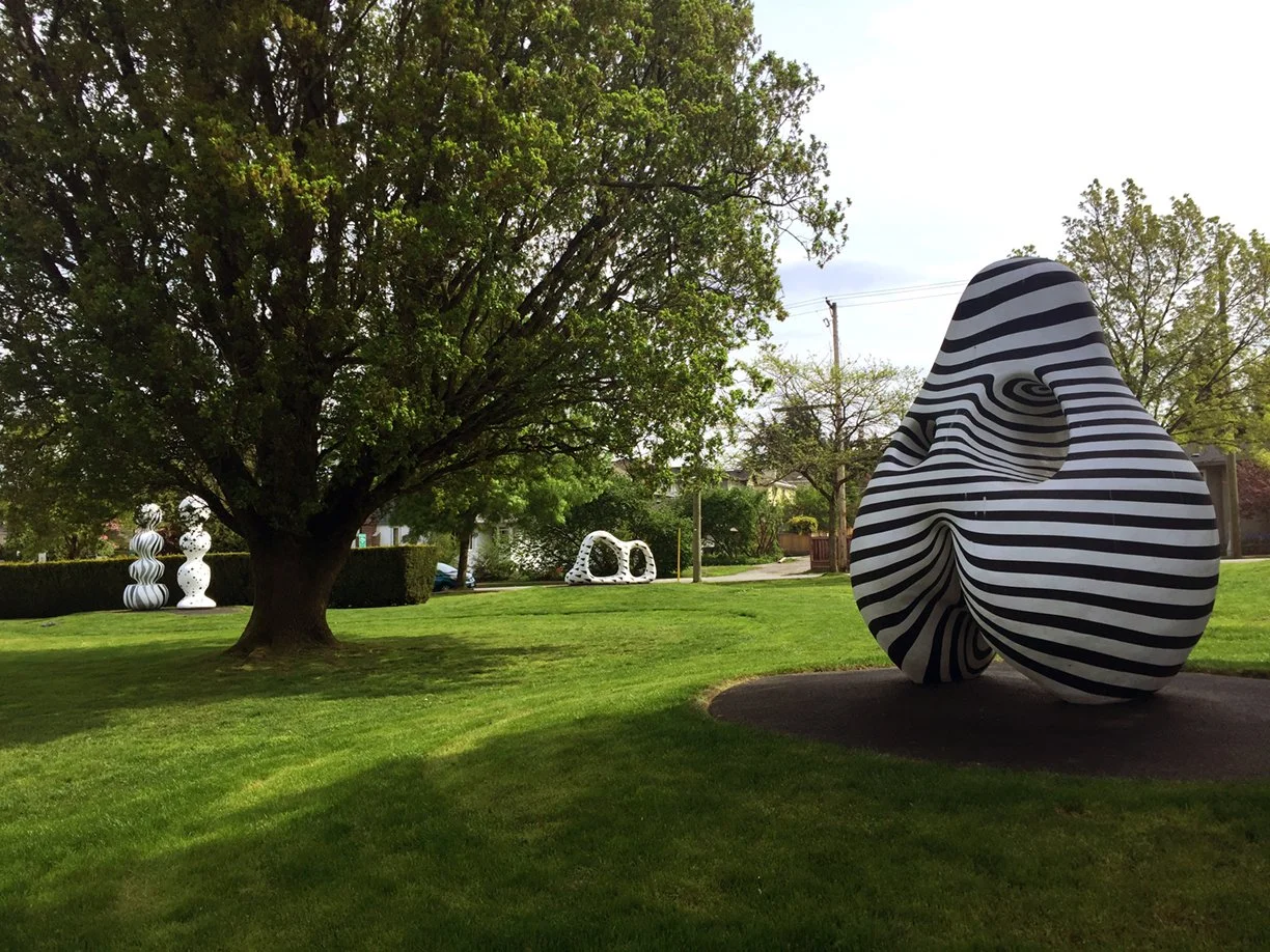 Outdoor park scene with green grass, a large leafy tree, and several abstract black and white striped sculptures.