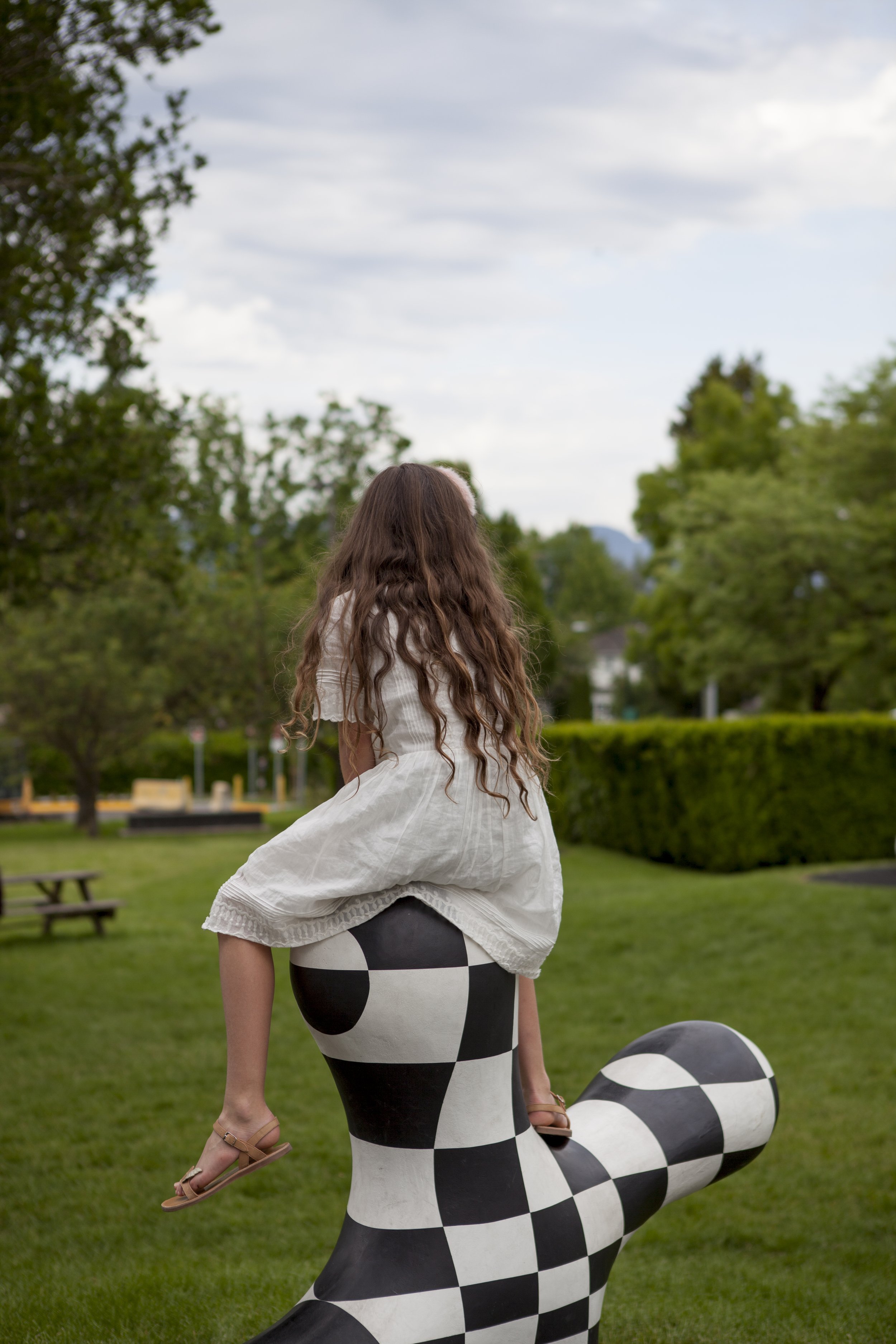 A woman with long wavy hair, wearing a white dress, is riding on the back of a sculpture of a black-and-white checkered snake in a park with trees, a bench, and cloudy sky in the background.