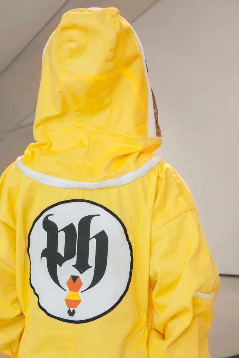 Back view of a person wearing a yellow jacket with a hood, featuring a large patch of the New York Yankees logo with a black letter 'h' and a stylized orange and black Y with a tie.