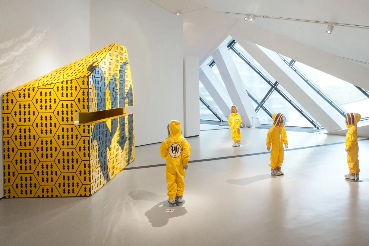 Children in yellow hazmat suits standing in an art gallery with modern architecture and large windows.