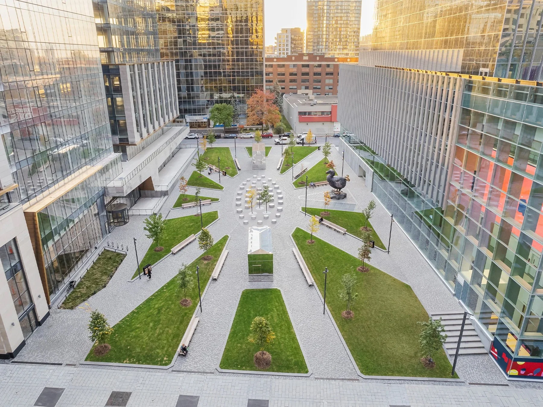 An aerial view of a modern cityscape showing a courtyard with green grass, trees with fall foliage, benches, walkways, and sculptures surrounded by tall glass office buildings.