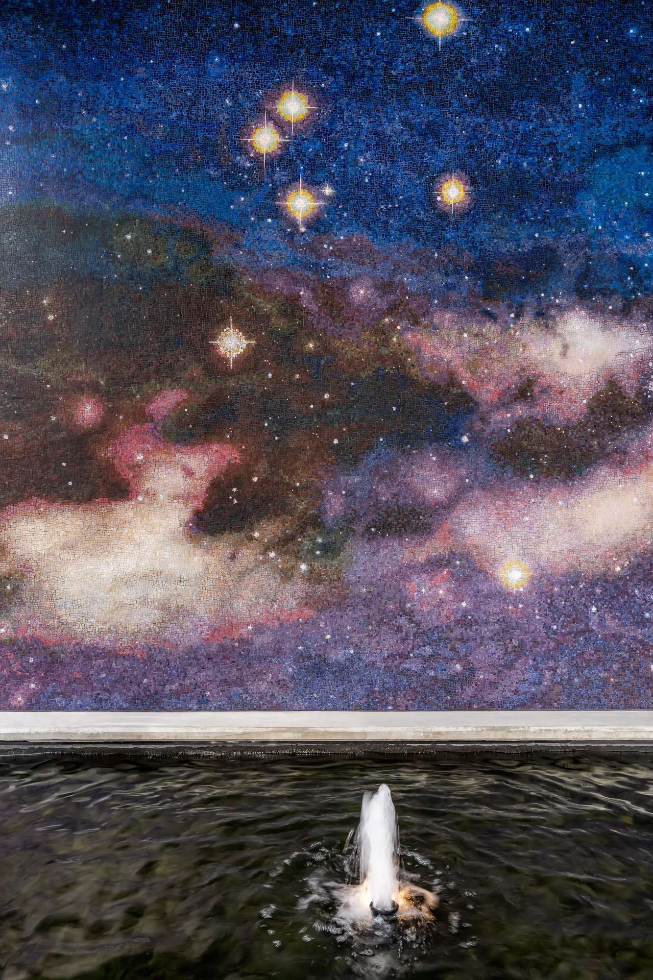 A fountain with water spraying upwards in the foreground, and a mosaic mural of a starry galaxy with bright stars and colorful nebulae on a wall in the background.