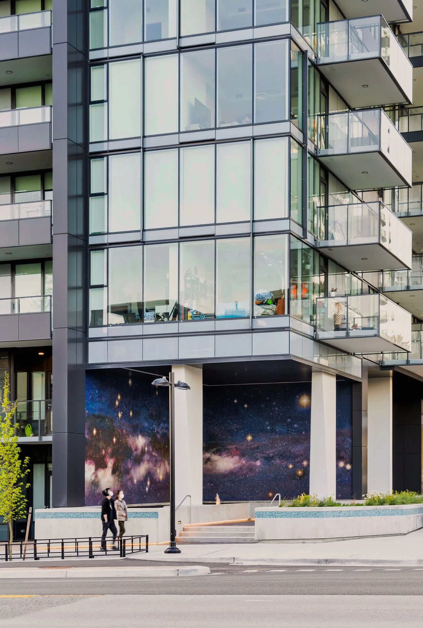 A modern multi-story building with glass balconies and a large mural of a galaxy on the lower facade. Two people are walking on the sidewalk in front of the building, and a street with a crosswalk and streetlights is visible in the foreground.