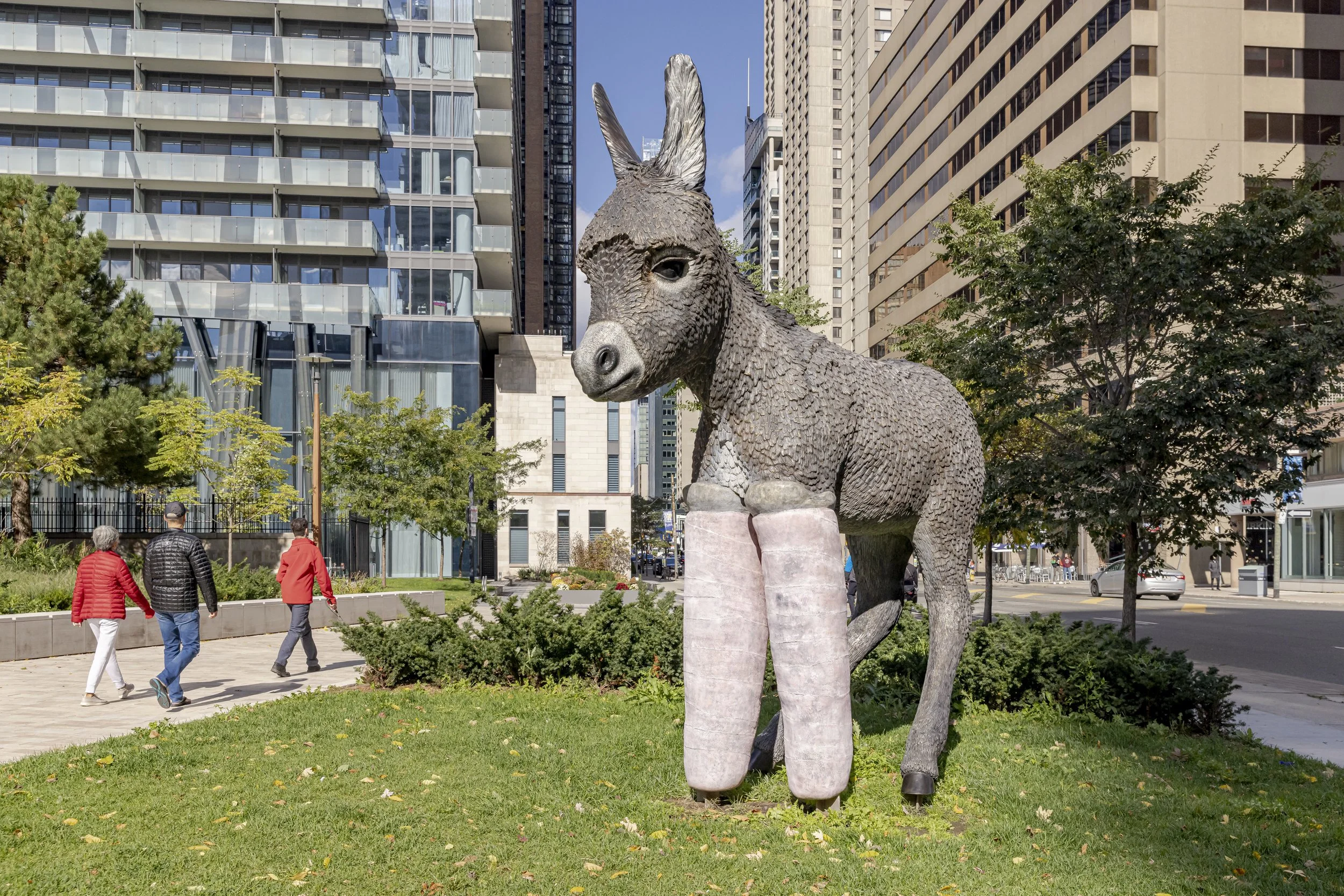 A large sculpture of a donkey made of textured materials stands in an urban park setting with tall modern buildings in the background. There are four people walking on the paved pathway nearby, and some trees and green bushes surround the area.