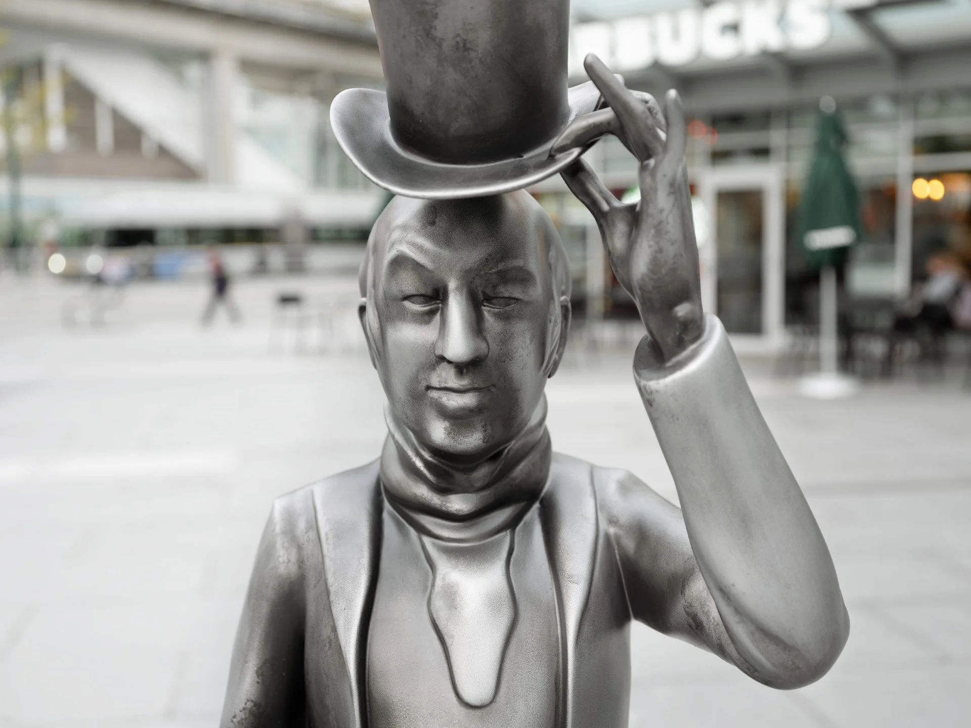 Close-up of a metallic sculpture of a man with a hat, holding a hat in his right hand, in an urban outdoor setting.