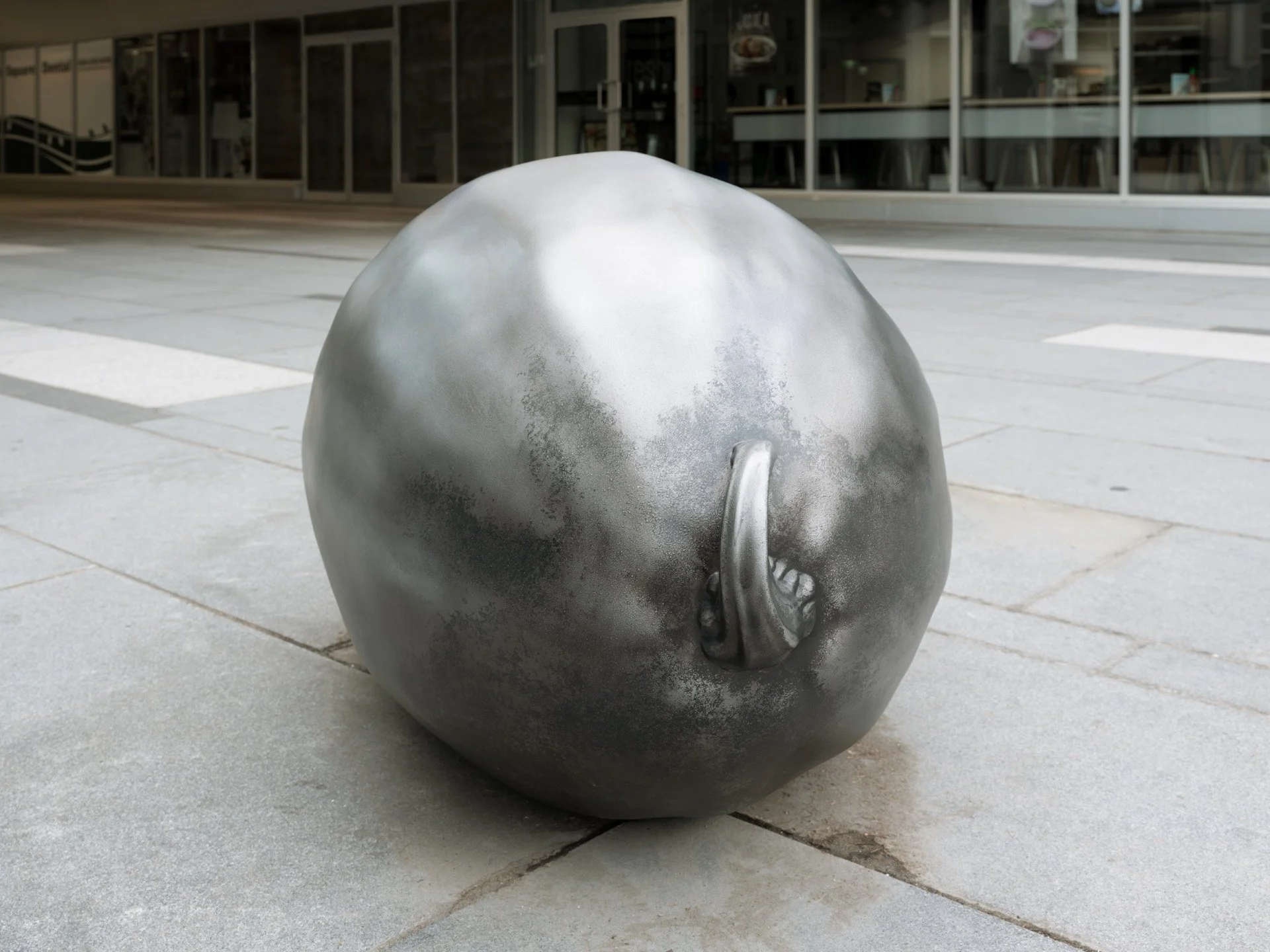 Sculpture of a round object with a zipper and a small ear, resembling a fruit or an abstract form, situated on a sidewalk in front of a building with glass windows.