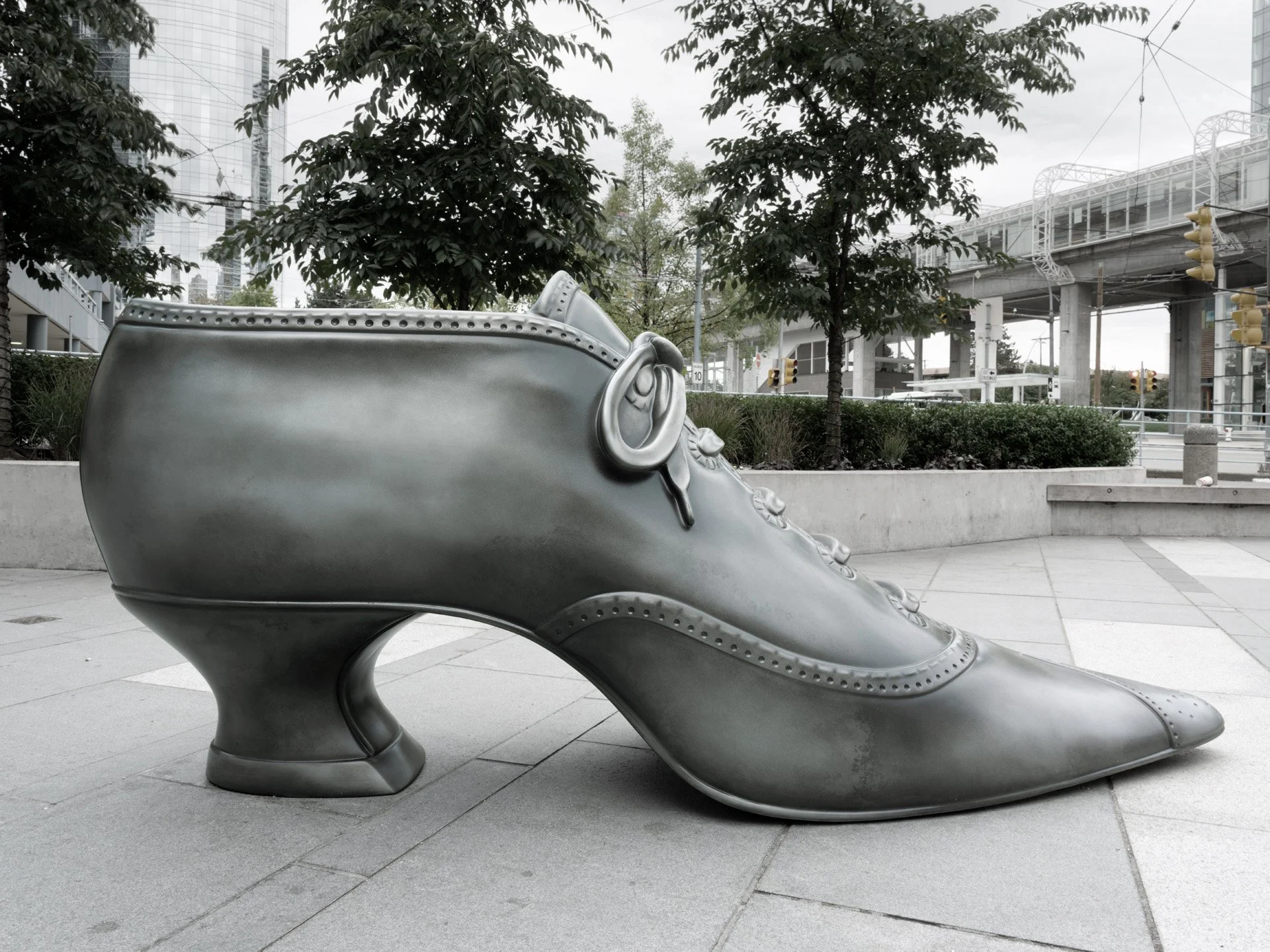 A large shiny silver shoe sculpture resembling a sneaker, situated outdoors on a paved area among trees and urban buildings in the background.