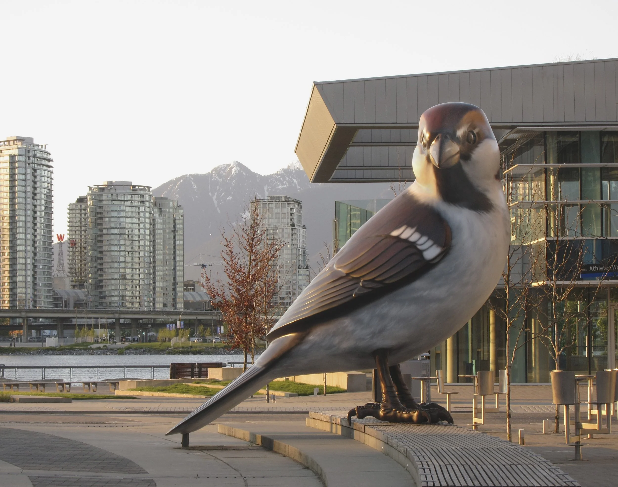 A large eagle sculpture made of metal is situated outdoors in front of modern buildings, with mountains in the background. The eagle appears to be perched on a ledge or platform.