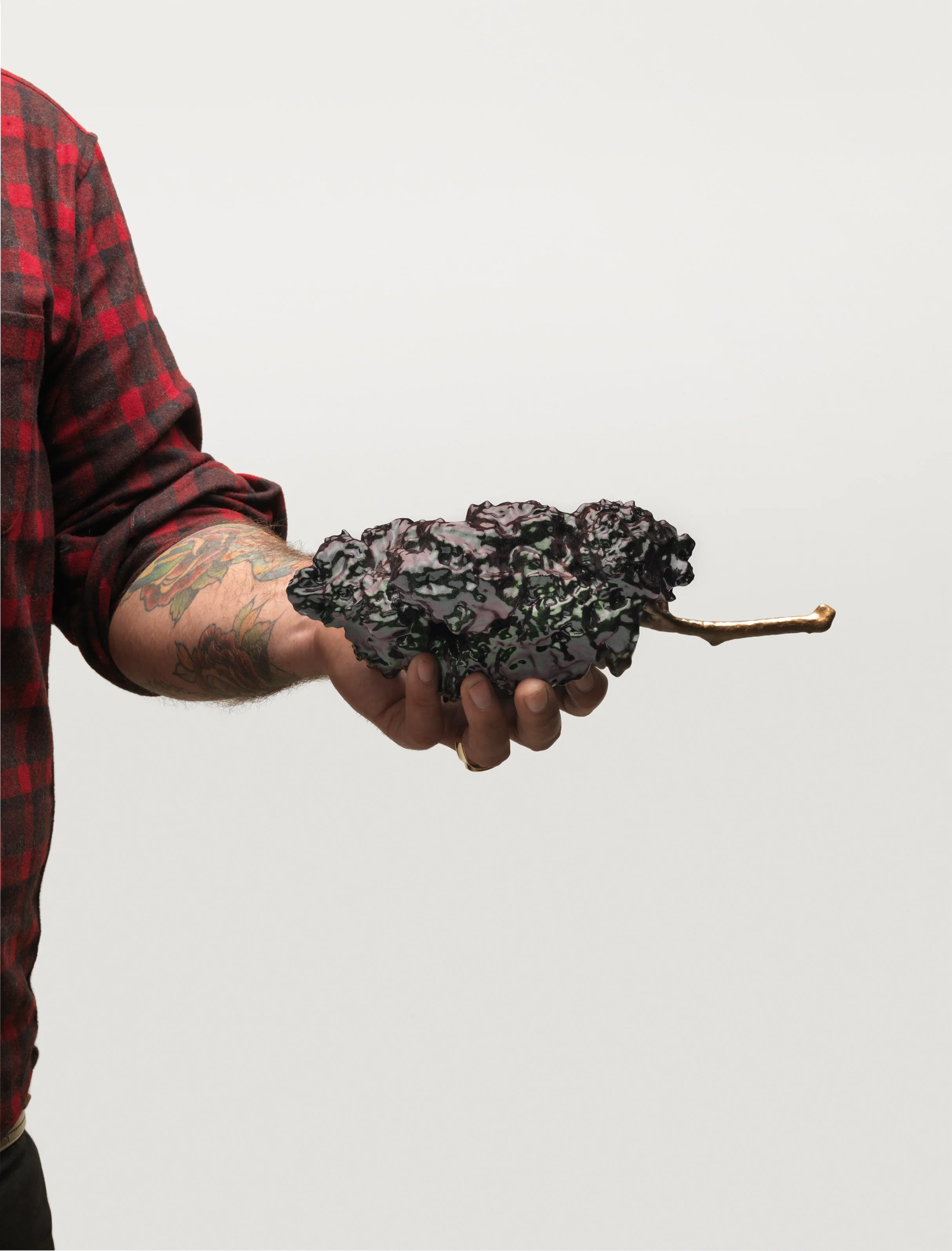 Person with tattooed arm wearing a red and black plaid shirt holding a black kale and purple marijuana bud ith a light gray background.