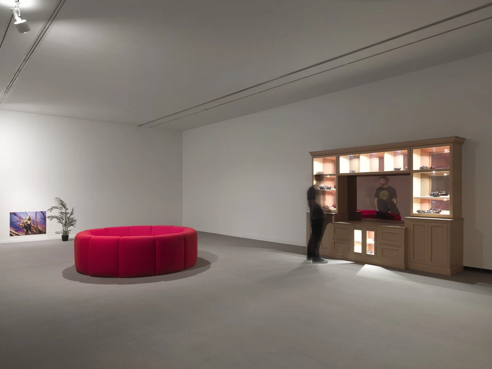 An art gallery with a red semi-circular sofa, a potted plant, and a wooden display cabinet with glass shelves and lighting. Two people are looking at the display, and there is a colorful painting on the wall.