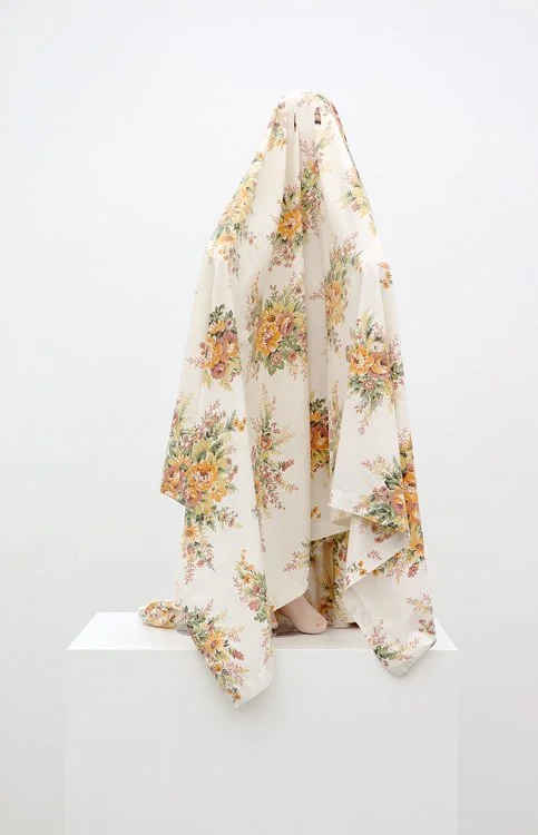 A floral fabric dress draped over a sculpture or mannequin, with only a small portion of a foot visible underneath, displayed against a plain white background.