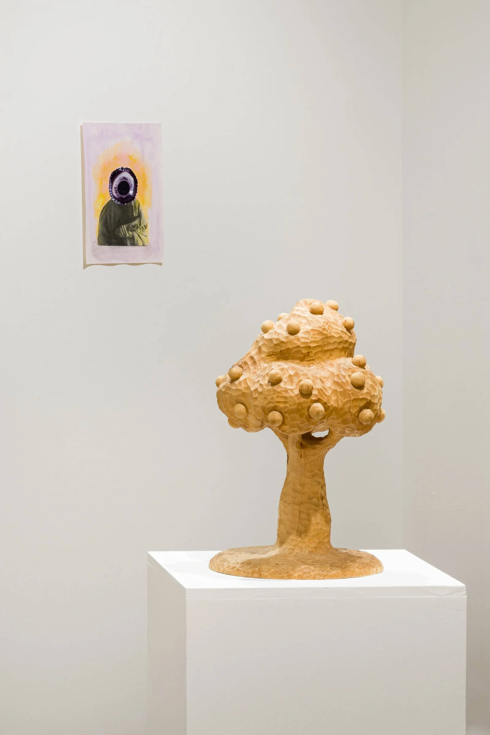 Wooden sculpture of a tree with a textured trunk and rounded top, displayed on a white pedestal in an art gallery, with a painted portrait of a person with a bird's eye on the face hanging on the wall behind it.