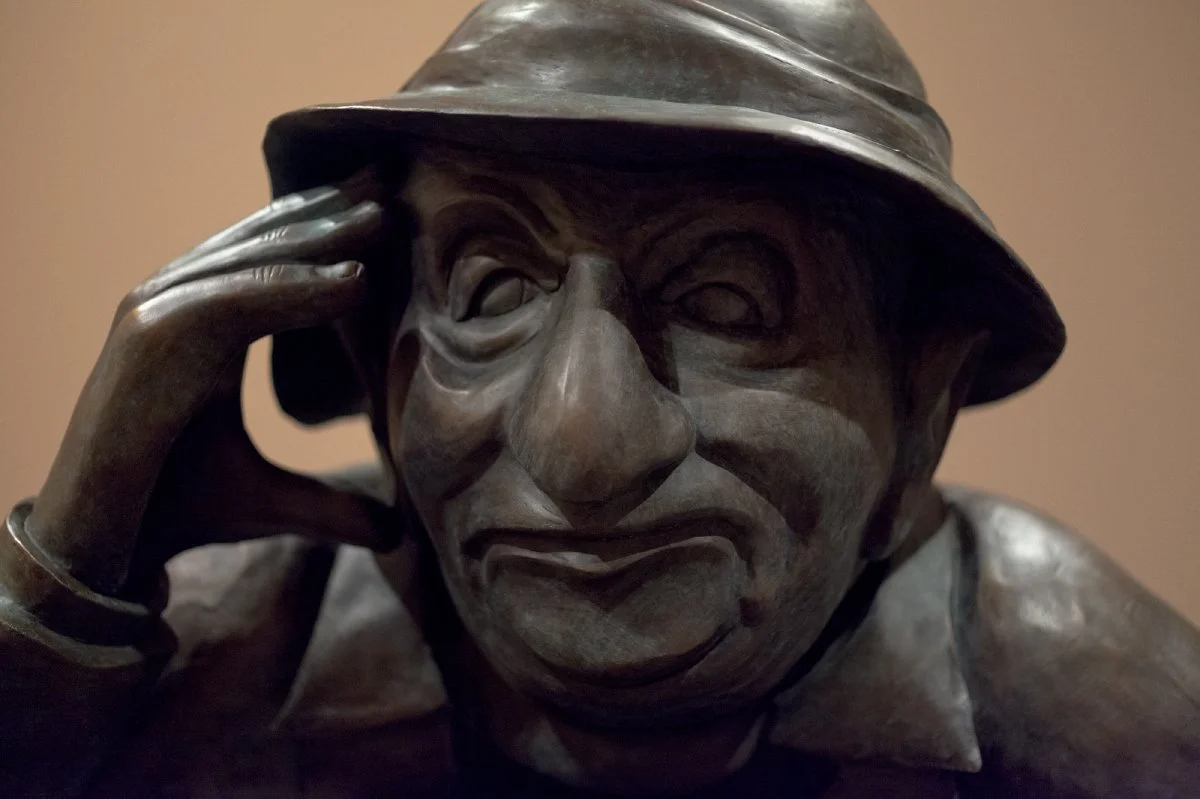 Bronze sculpture of a man with a hat rubbing his temple with his right hand.