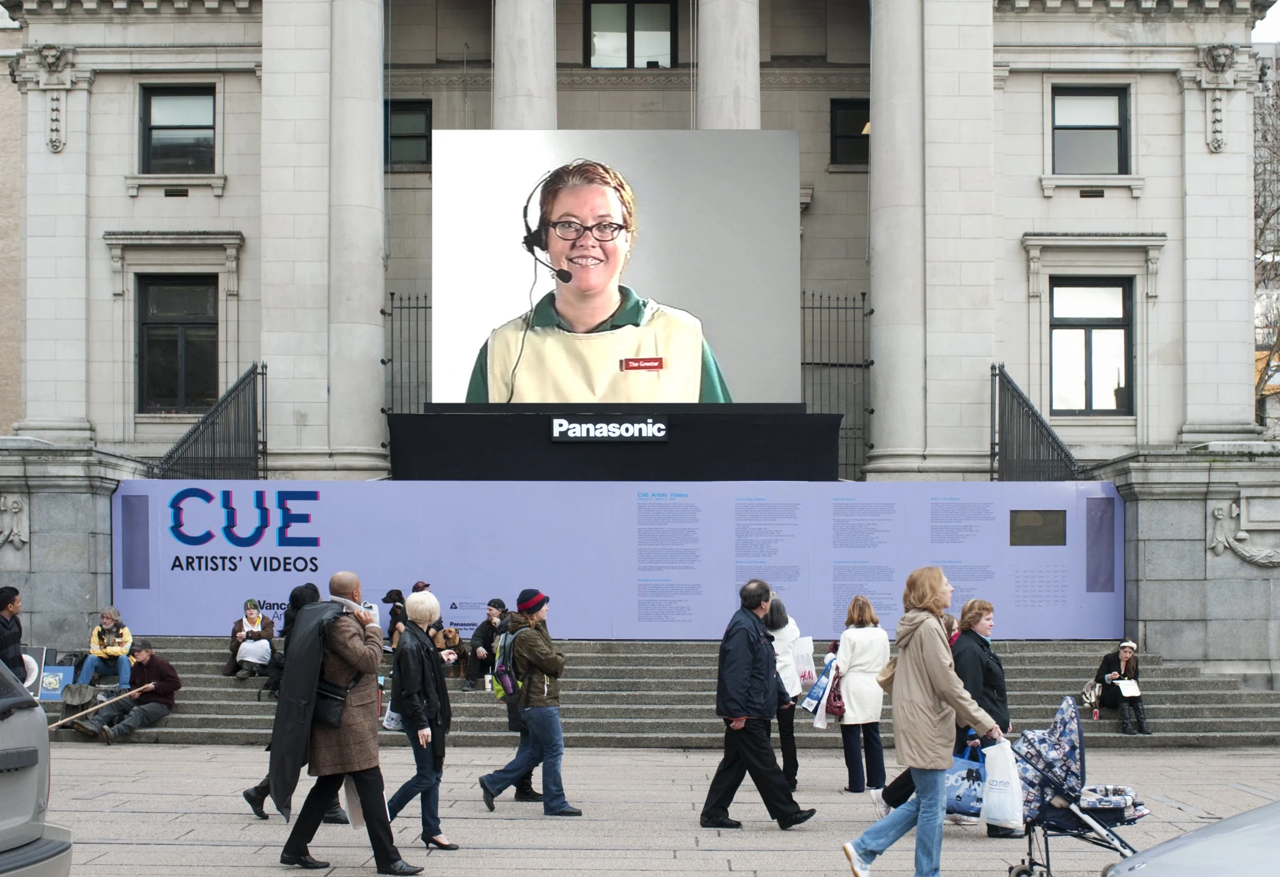 Outdoor event with a large digital screen displaying a smiling woman with glasses and a headset, set against a historic building with columns. Several people are walking and sitting on the steps in front of the screen, some holding bags, with a banner that reads "CUE Artists' Videos" and other informational posters.