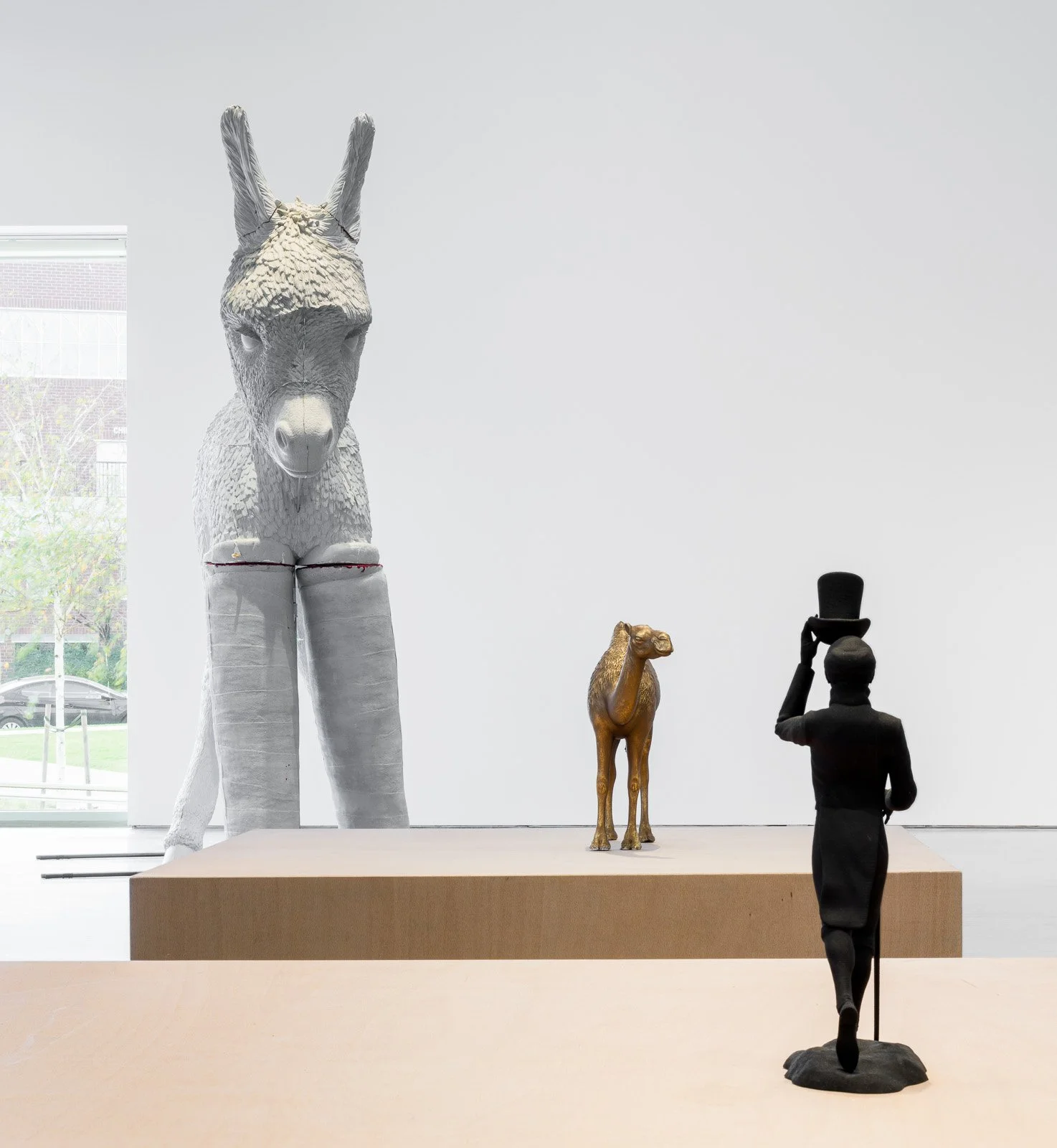 Gallery with animal sculptures, including a donkey and a camel, and a person sculpture holding a top hat, all displayed on a platform near a window.
