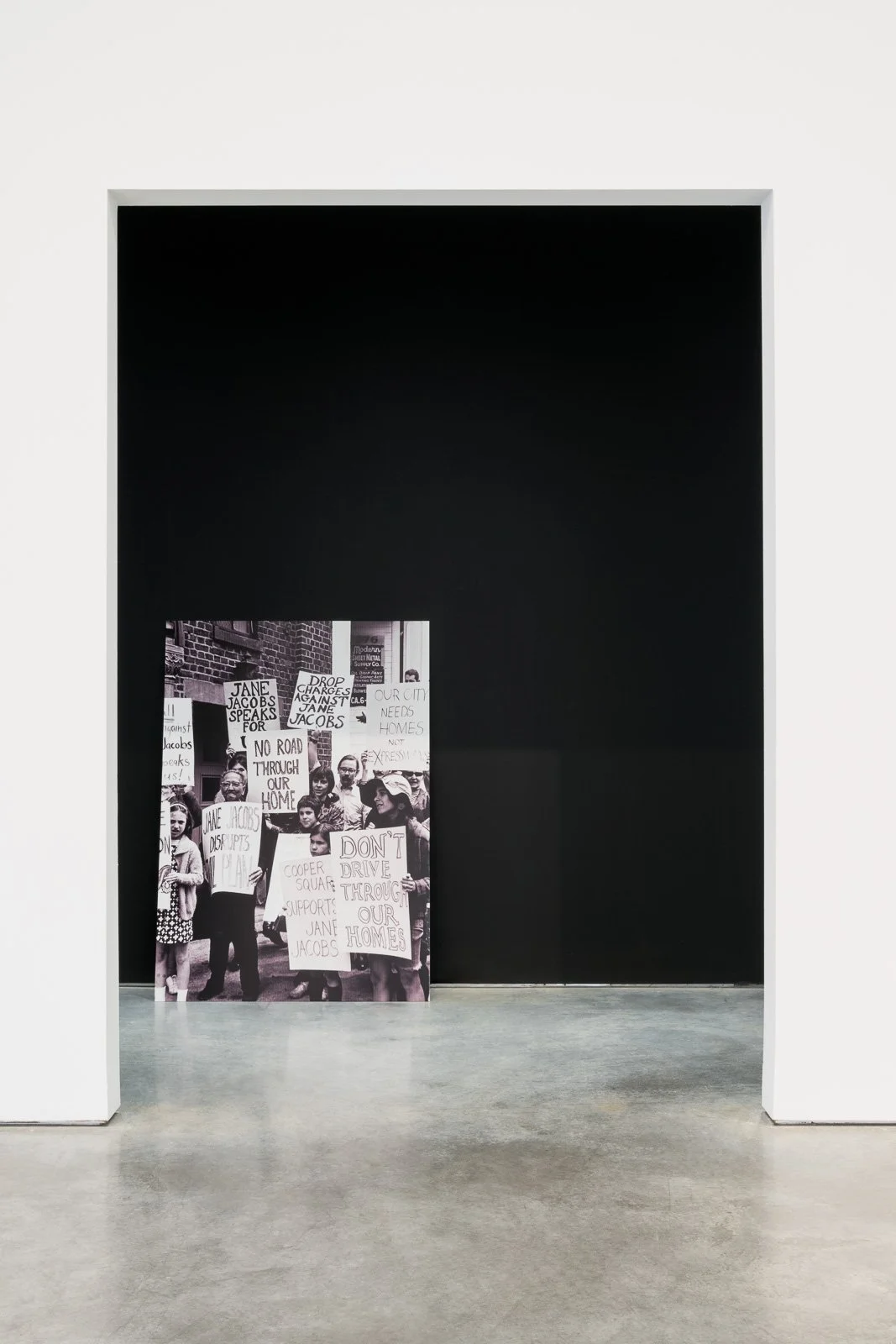 A black and white photograph of a protest with people holding signs that read, 'Jane Jacobs Speaks for Us!', 'Drop Charges Against Jane Jacobs', 'No Road Through Our Home', 'Your City Needs Homes Not Xpress', and 'Don't Drive Through Our Homes', displayed on an easel in front of a black wall.