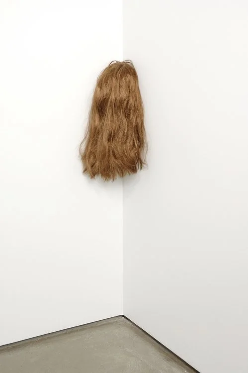 A wig with long, wavy, brown hair hanging on a white wall in a corner of a room.