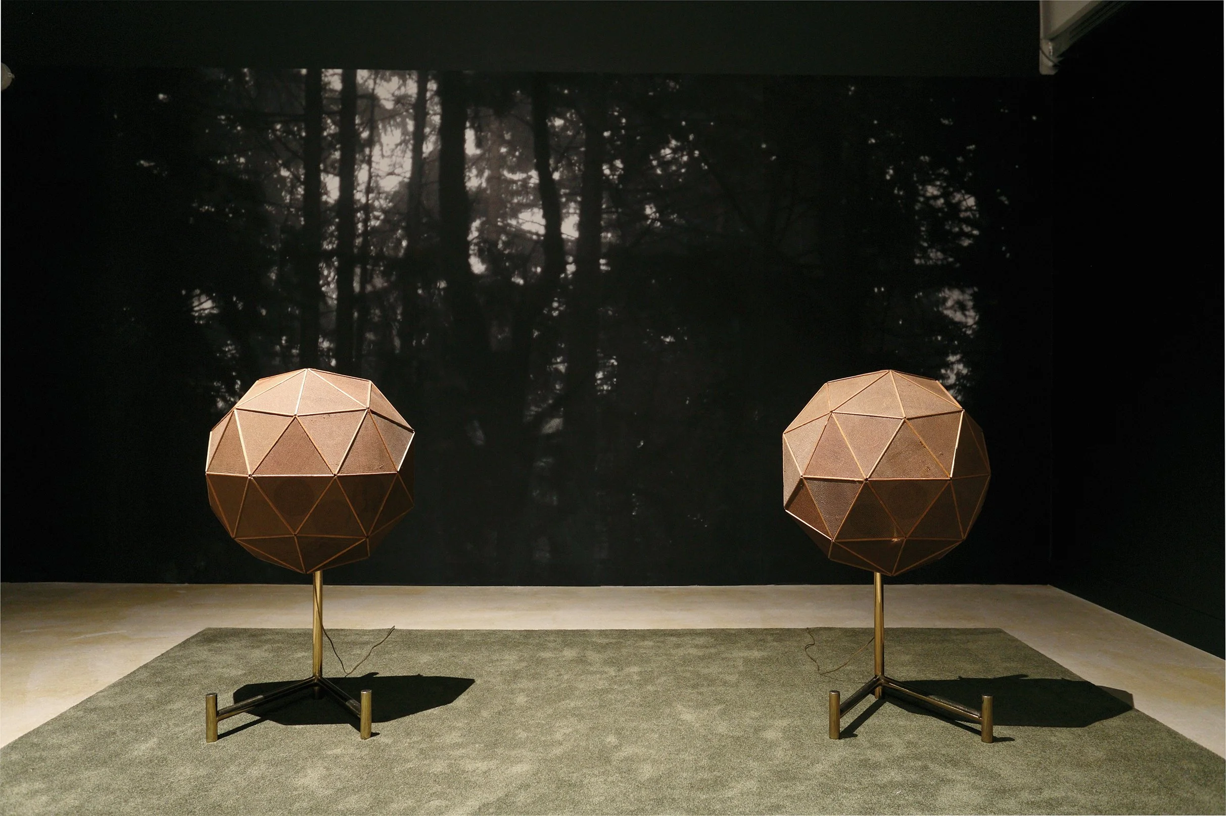 Two tan-coloured, geodesic speakers stand on a stage with a dark backdrop showing trees and the night sky.