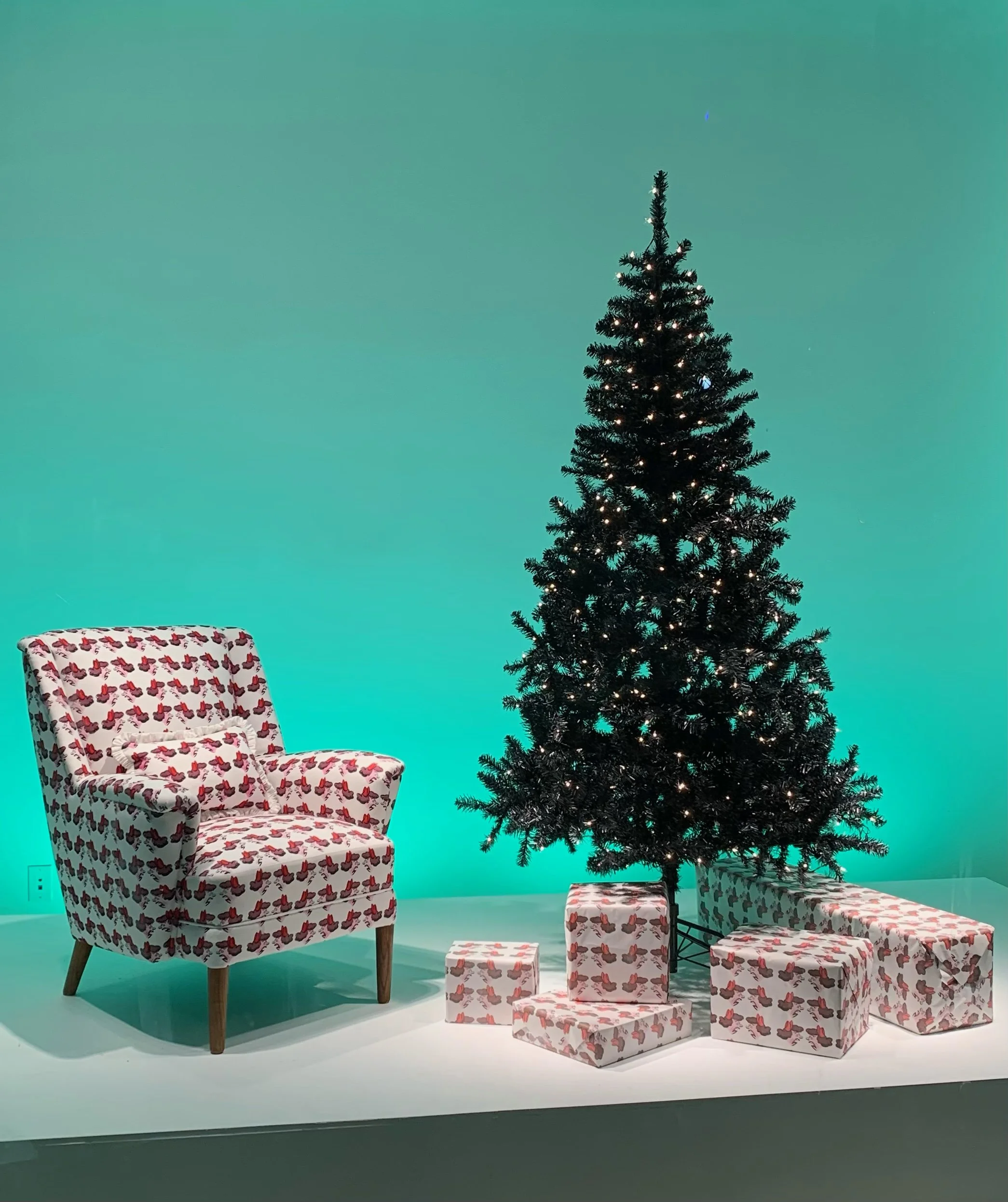 Display of Christmas decorations including a tall Christmas tree with small white lights, a patterned armchair with a middle-aged woman and several wrapped presents in paper with, set against a aqua background.