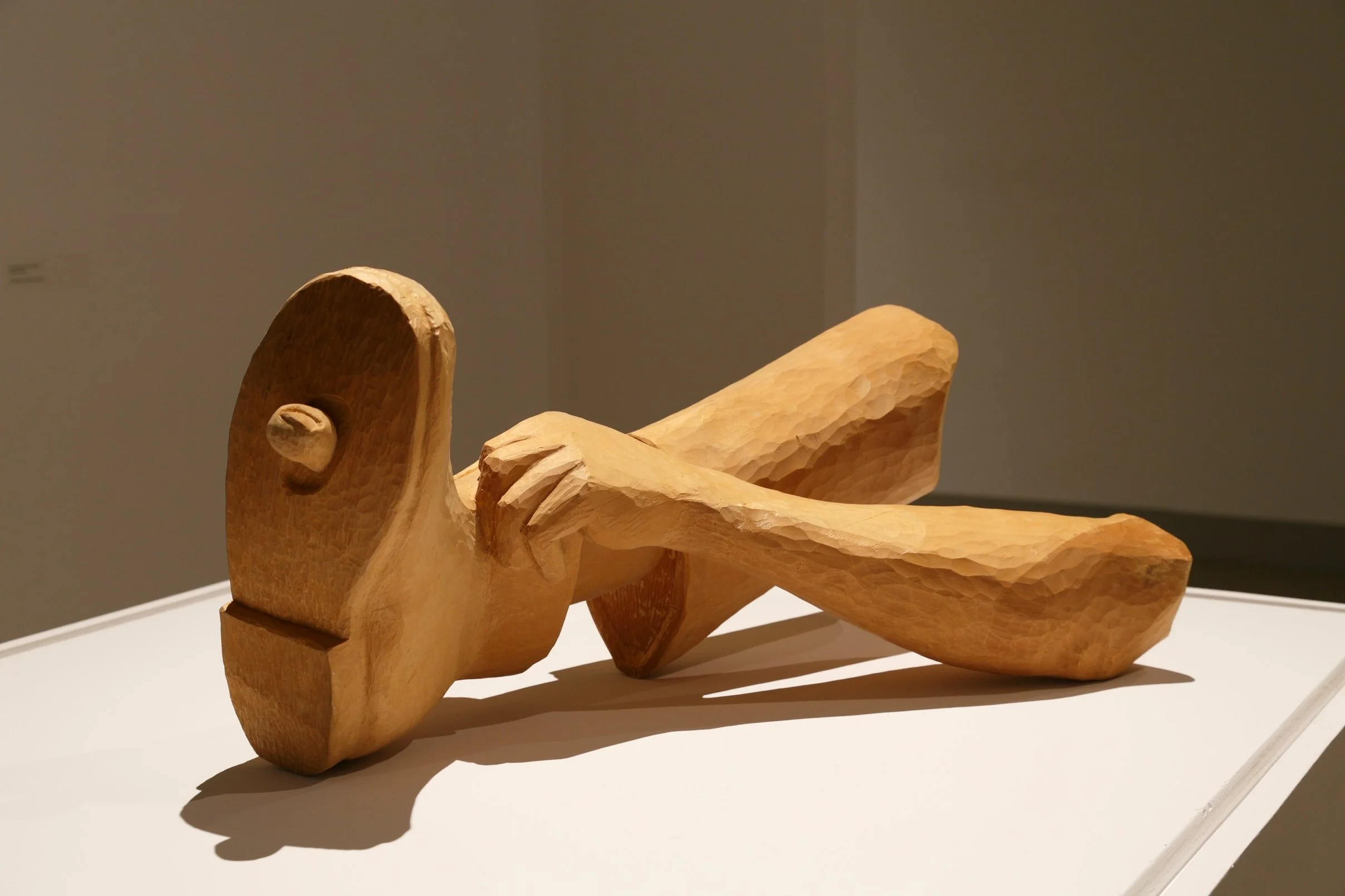 Wooden sculpture of a reclining figure with a smooth head and carved limbs, displayed on a white pedestal.