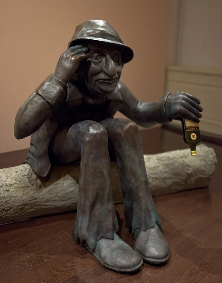 Bronze sculpture of a man sitting on a log, wearing a hat and jacket, holding a small bottle in his right hand, with a thoughtful expression.