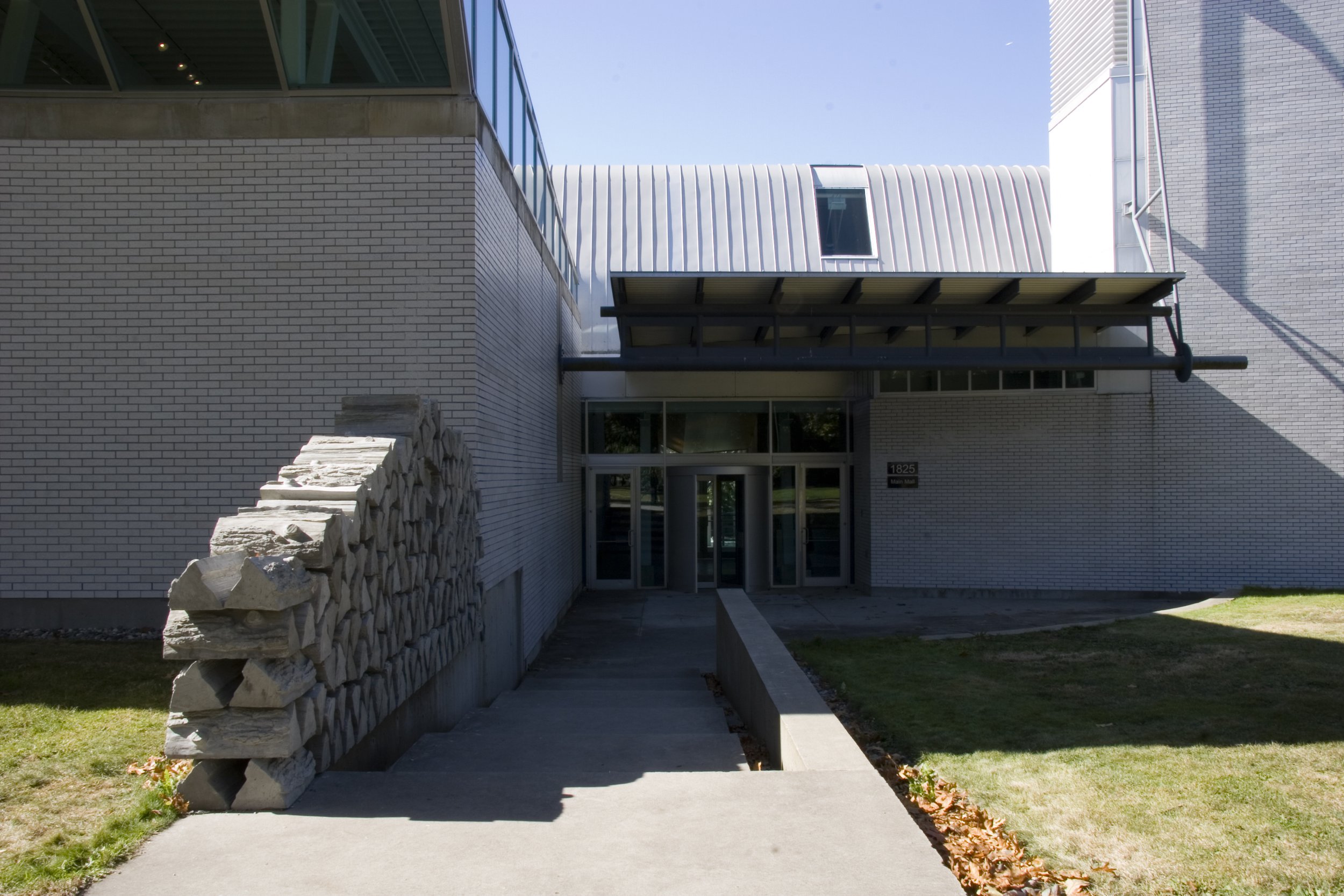 Modern building exterior with brick walls, large glass windows, and a metal roof. Stone wall and sidewalk leading to entrance.