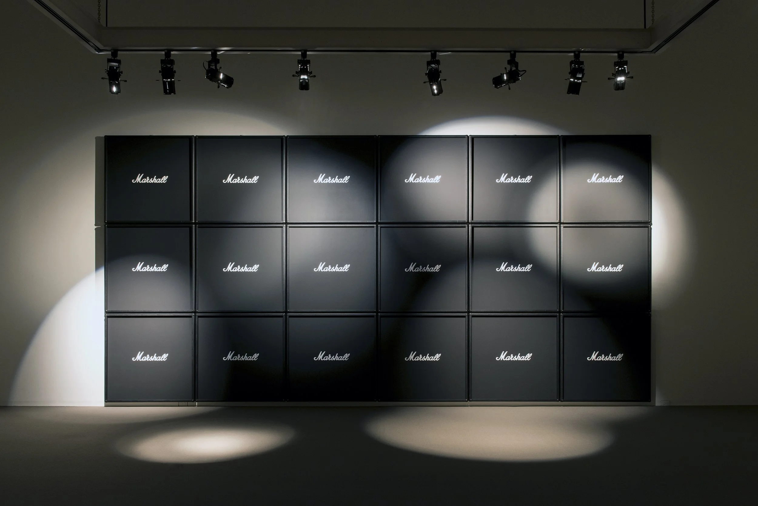 Black Marshall speaker cabinets stacked in a grid formation with overhead spotlights illuminating them in a dark room.