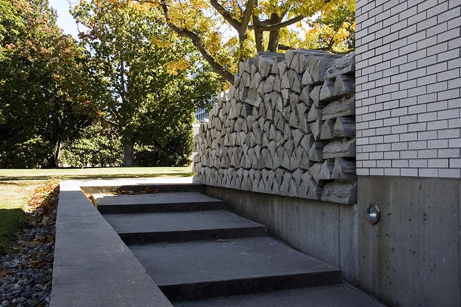 Concrete stairs leading up beside a white brick wall with stacked firewood against it, surrounded by trees with fall foliage.