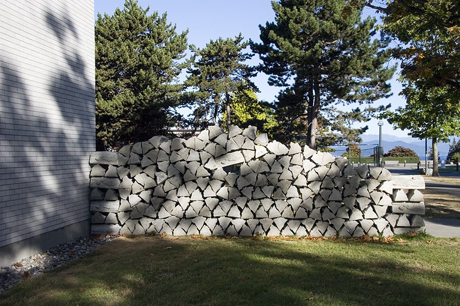 A neatly stacked pile of firewood logs placed against the side of a building. The background shows tall evergreen trees, a park bench, and a clear blue sky.