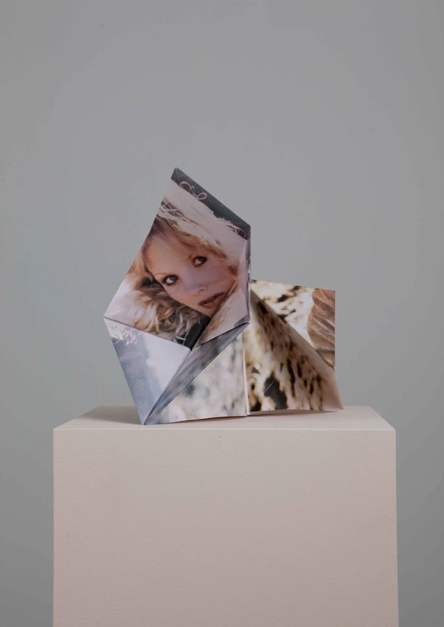 Contemporary art origami sculpture made of folded magazine pages with a woman's face, displayed on a white pedestal against a plain grey background.