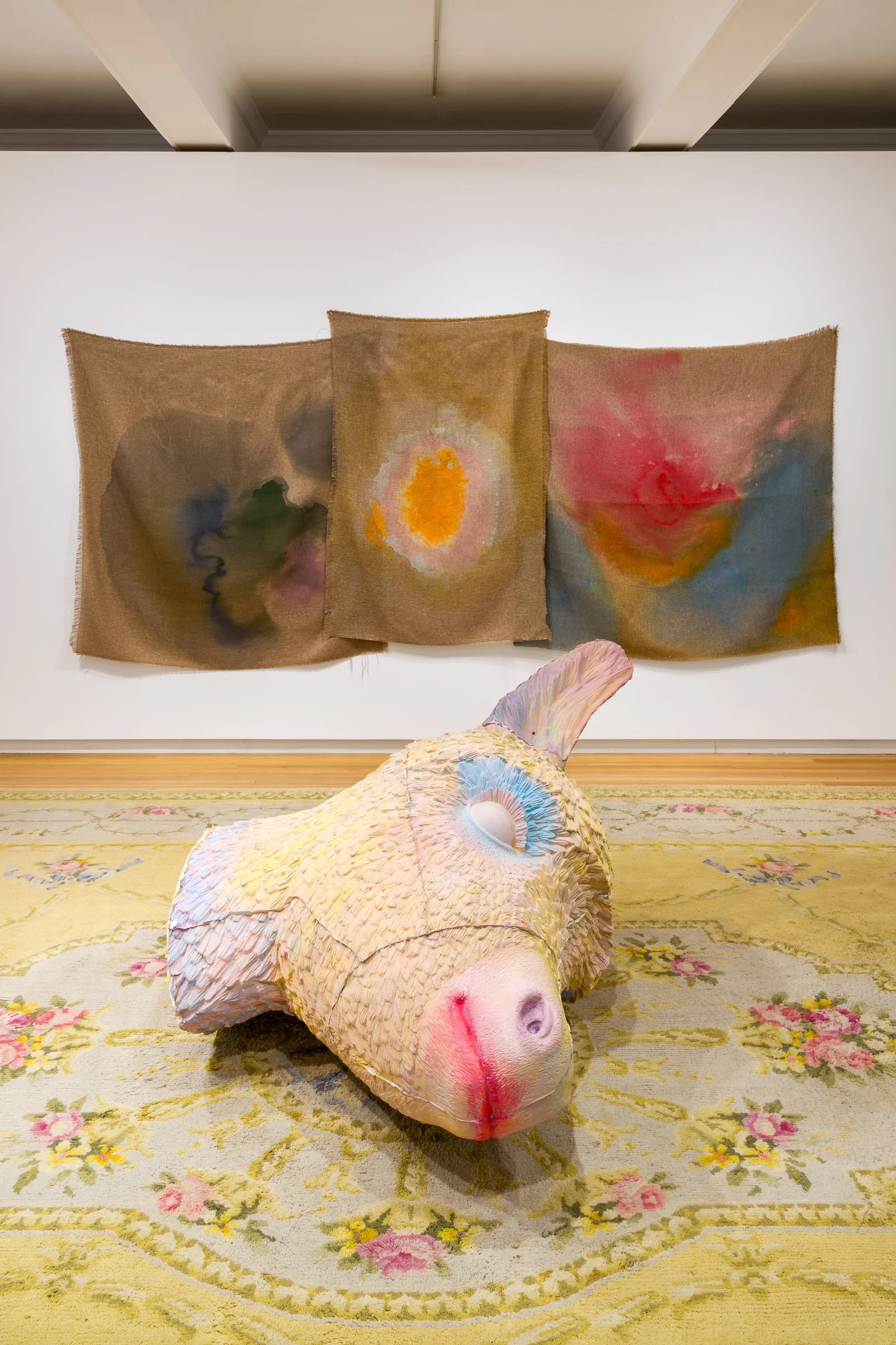 An art installation featuring a sculpture of a donkey's head with a textured, iridescent surface resembling shell material, placed on a floral-patterned rug. In the background, three colour-stained fabric panels on a wall.