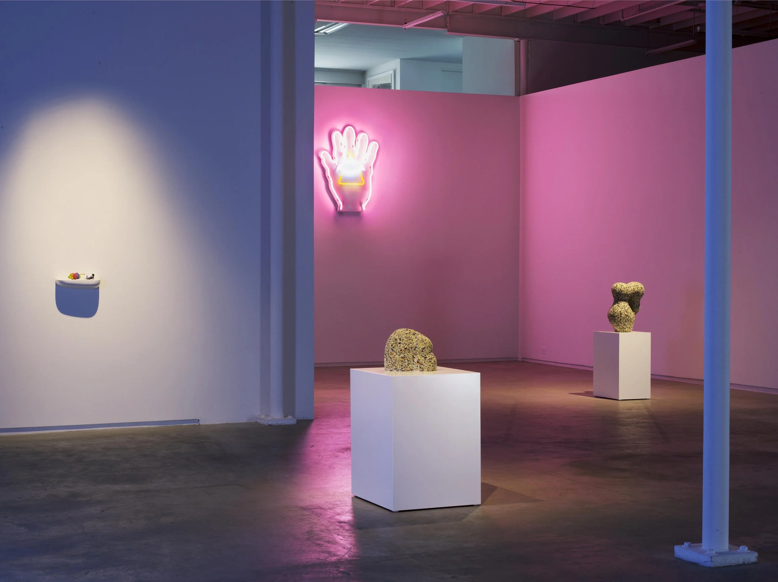 An art gallery with sculptures on white pedestals, colorful lighting, and a neon sign of a hand with a ring on the pink wall.