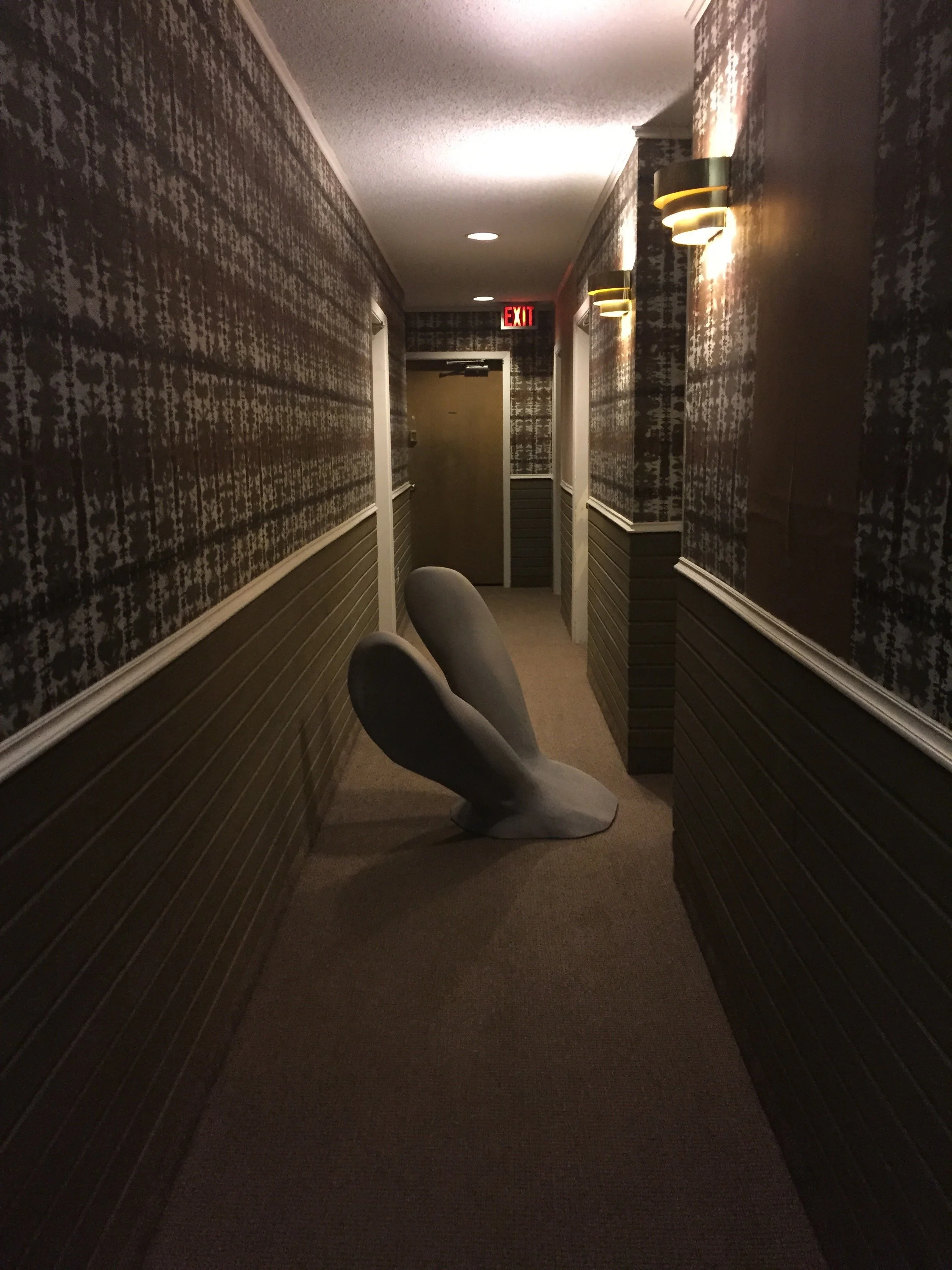 Hallway with patterned wallpaper, a pair of large grey rabbit ears, and a closed door at the end, illuminated by wall-mounted lights and ceiling lights.