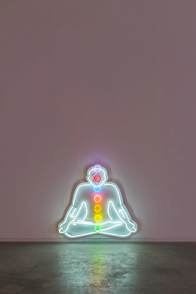 Neon art of a person meditating seated in a cross-legged position, with colored circles representing chakras aligned along the body from head to pelvis, against a plain background.