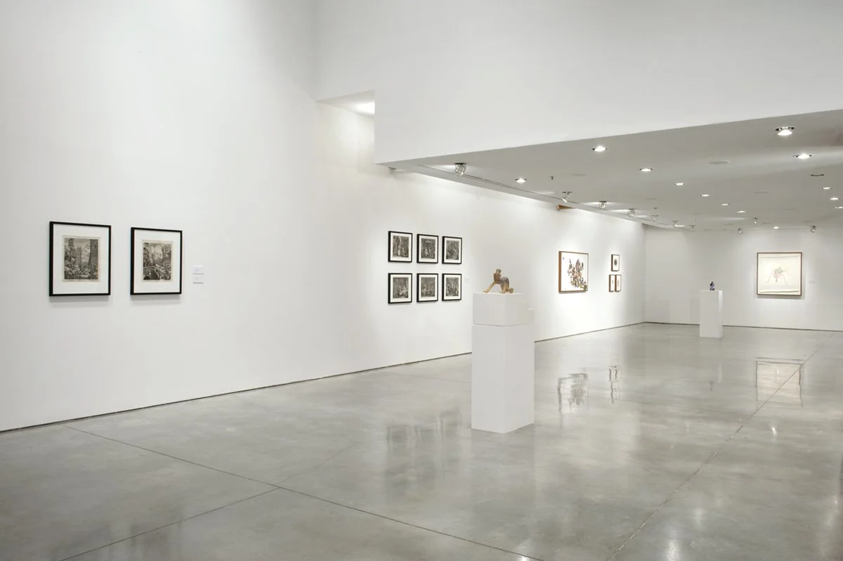 Empty art gallery with framed artwork on walls and sculptures on white pedestals.