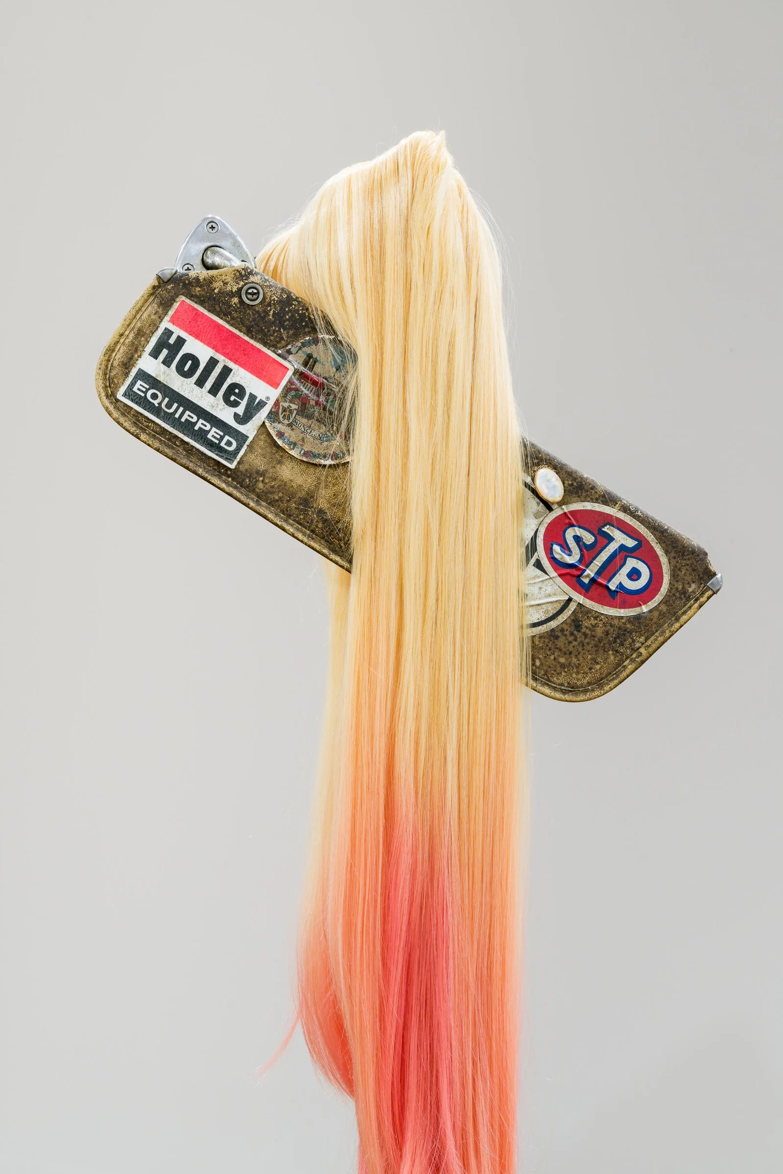 An old car visor with stickers, including a 'Holley Equipped' sticker and a 'STOP' sticker, holding a long, straight, pink-colored synthetic wig.