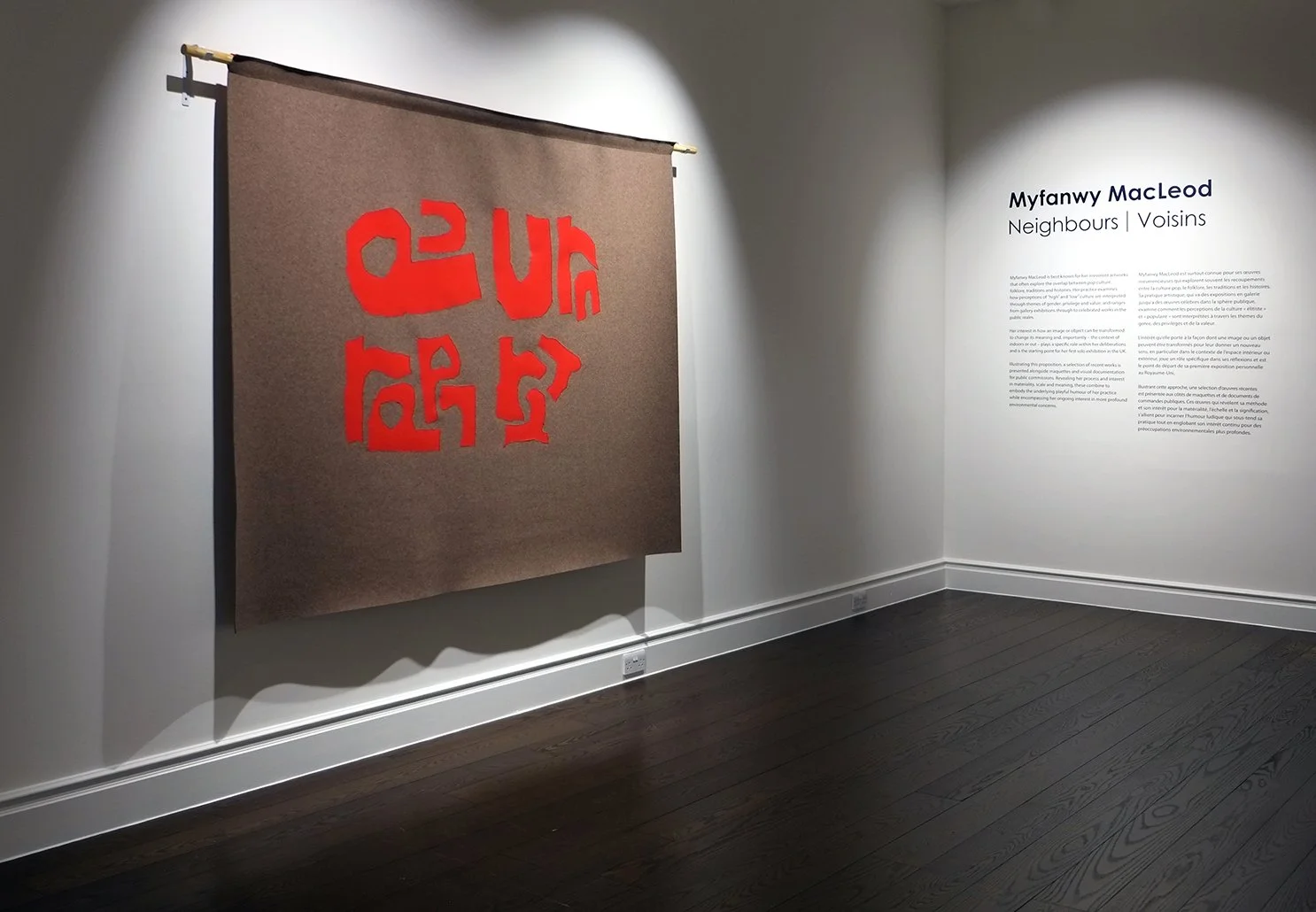 Art exhibit featuring a large brown felt banner with red abstract jumbled letters that reads purr, purr, purr, and a wall with text titled 'Myfanwy MacLeod Neighbours Voisins' on the right.