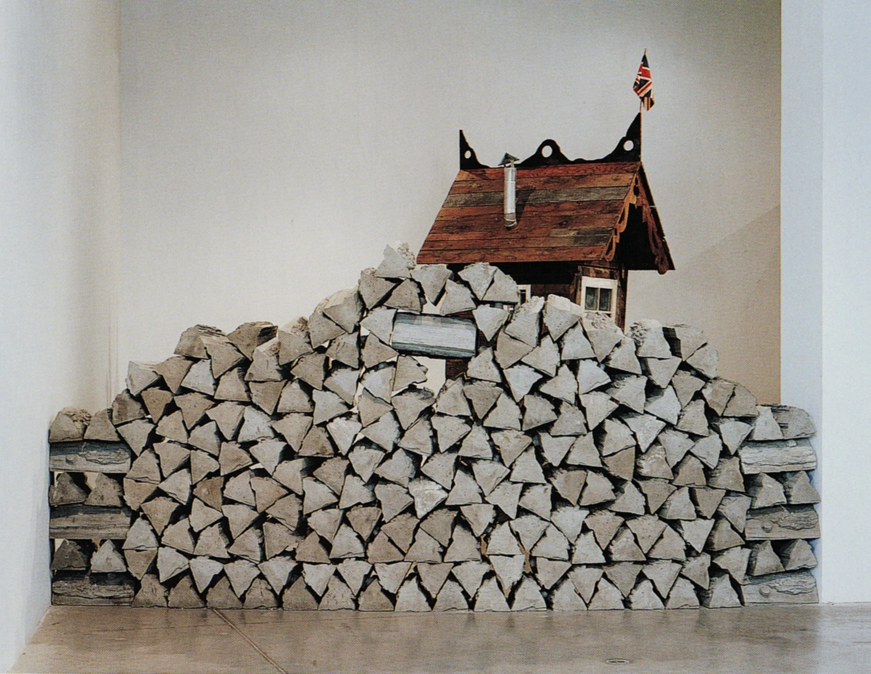 A large stack of chopped firewood arranged neatly against a white wall with a small wooden house model on top. The model has a small window, a chimney, and a Union Jack flag.