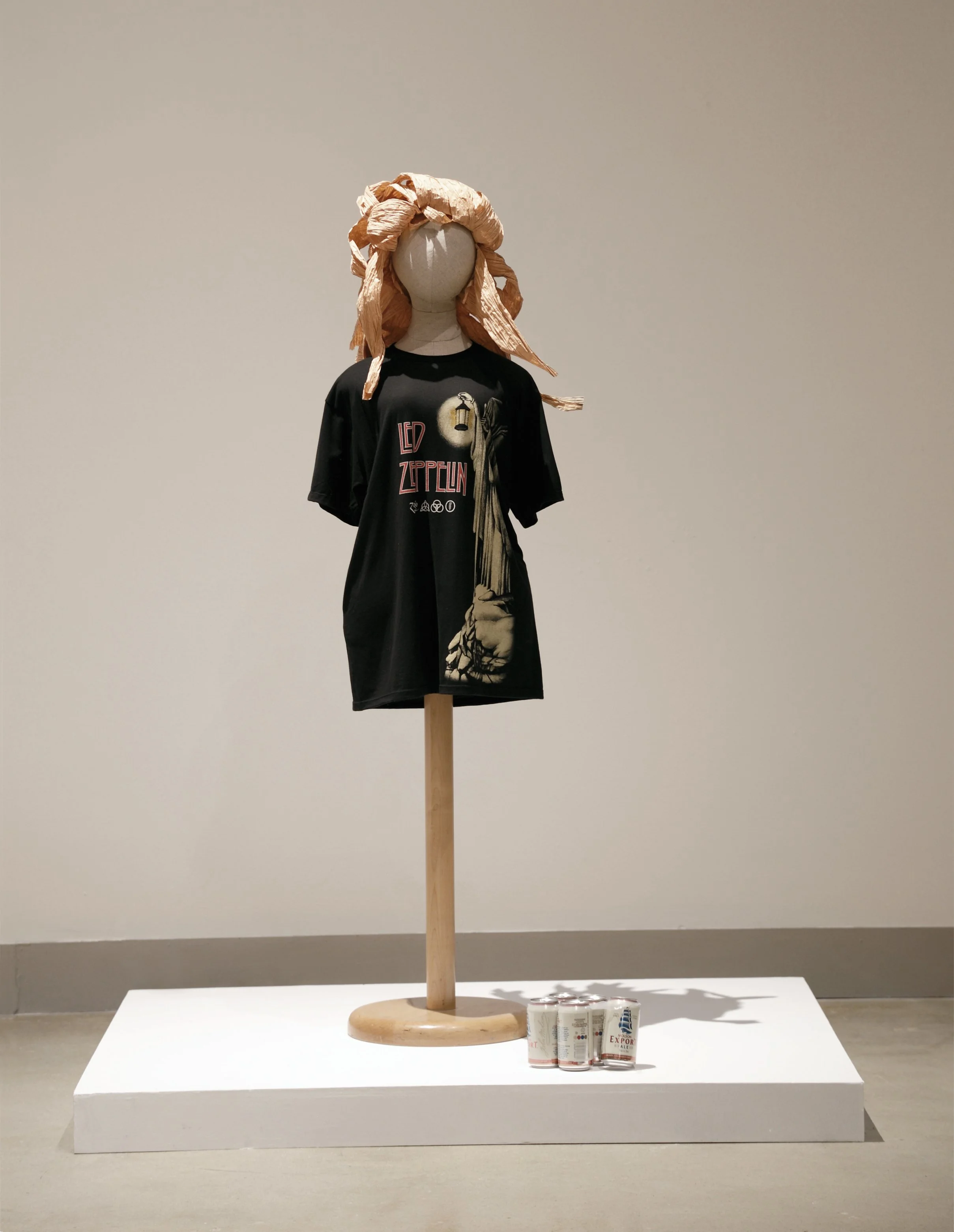 Mannequin with a beige paper head and hair, wearing a black Led Zeppelin T-shirt, standing on a wooden stand, displayed on a white platform with beer cans in front, against a plain gray wall.