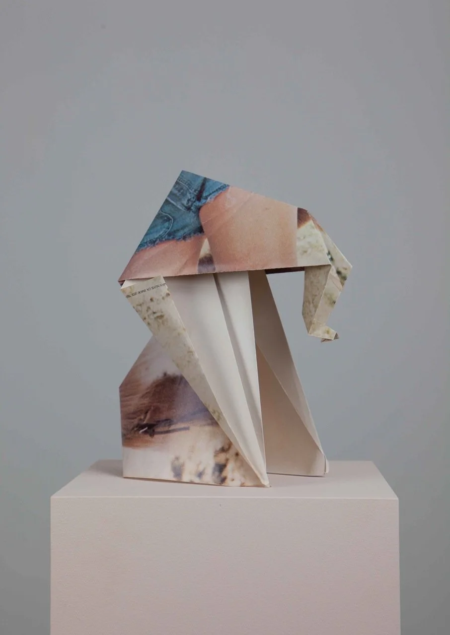 An abstract origami sculpture made from folded and cut magazine pages, shaped into a geometric form, displayed on a white pedestal against a gray background.