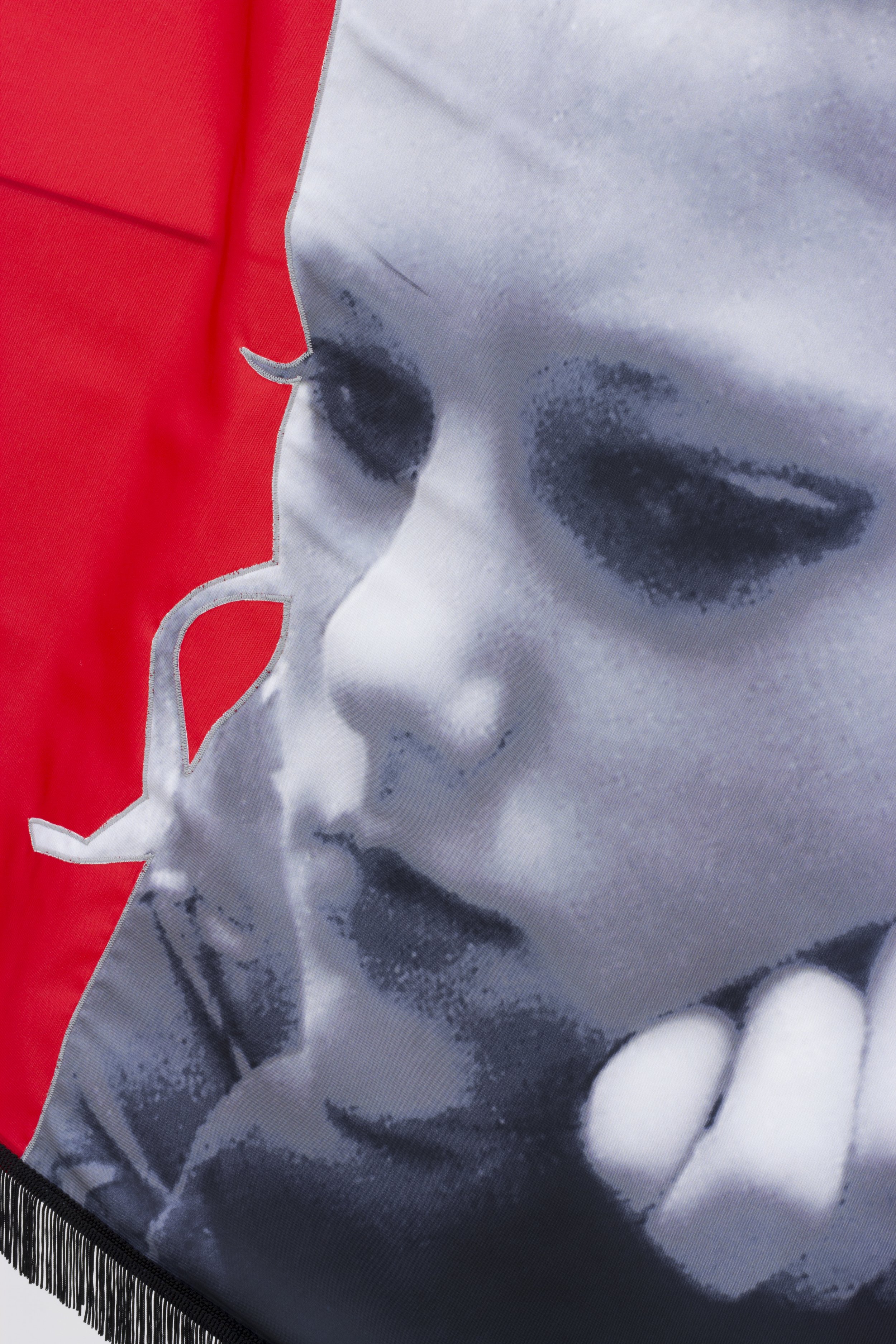 Close-up of a fabric with a black-and-white printed image of a girl's face, partially covered by a red-shaped overlay, and detailed with black fringes at the bottom.
