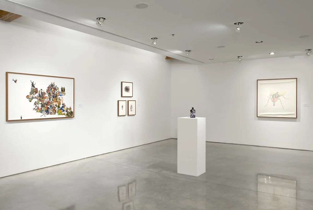 Contemporary art gallery with various framed artworks on white walls and a small sculpture on a white pedestal in the center of the room.
