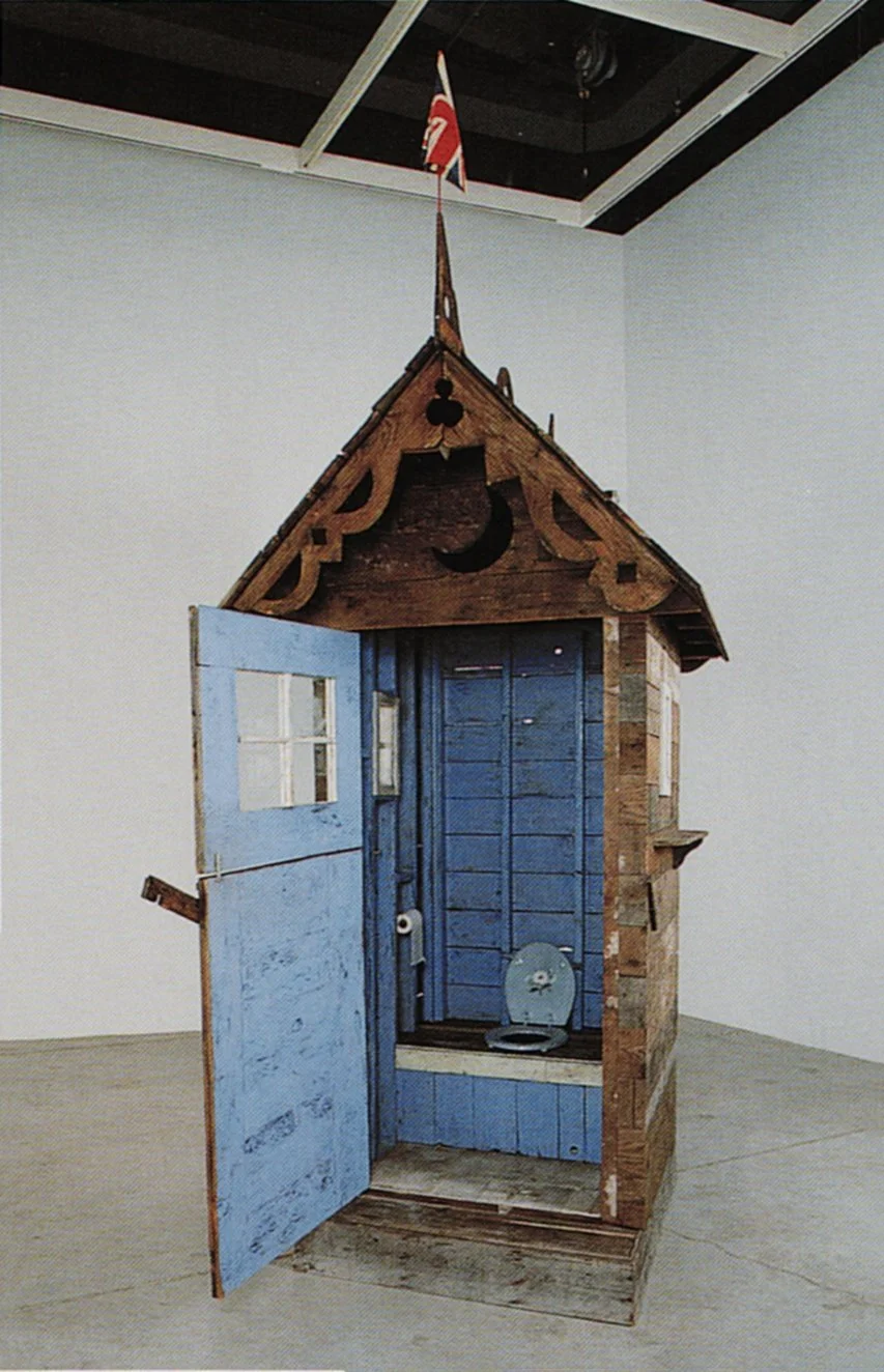 An art installation resembling a small wooden house converted into a quirky toilet, painted blue with a toilet seat inside, in a gallery space.