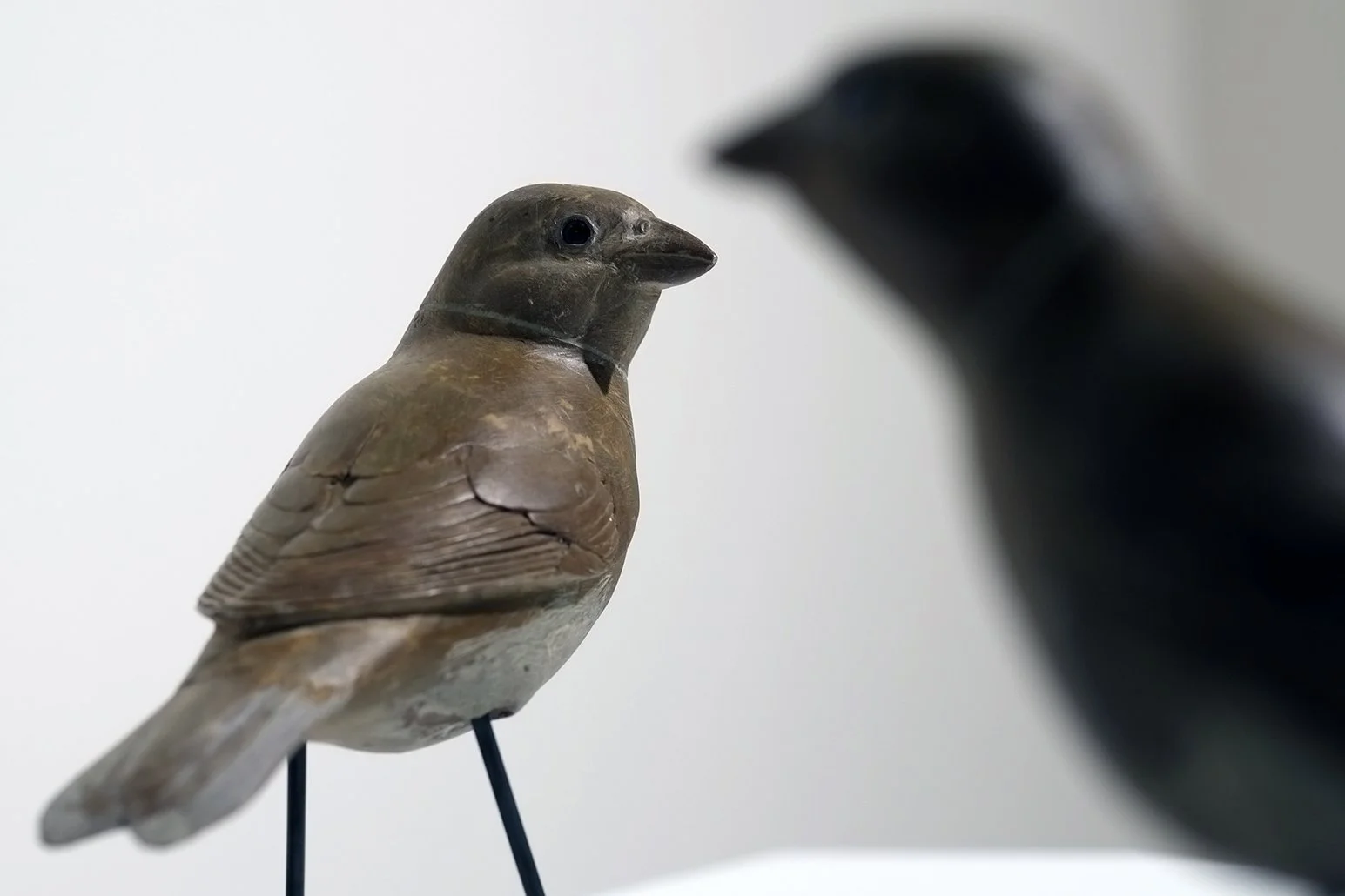A wooden bird sculpture with a detailed carved face and textured wings, mounted on a stand, with a blurred dark-colored bird sculpture in the background.