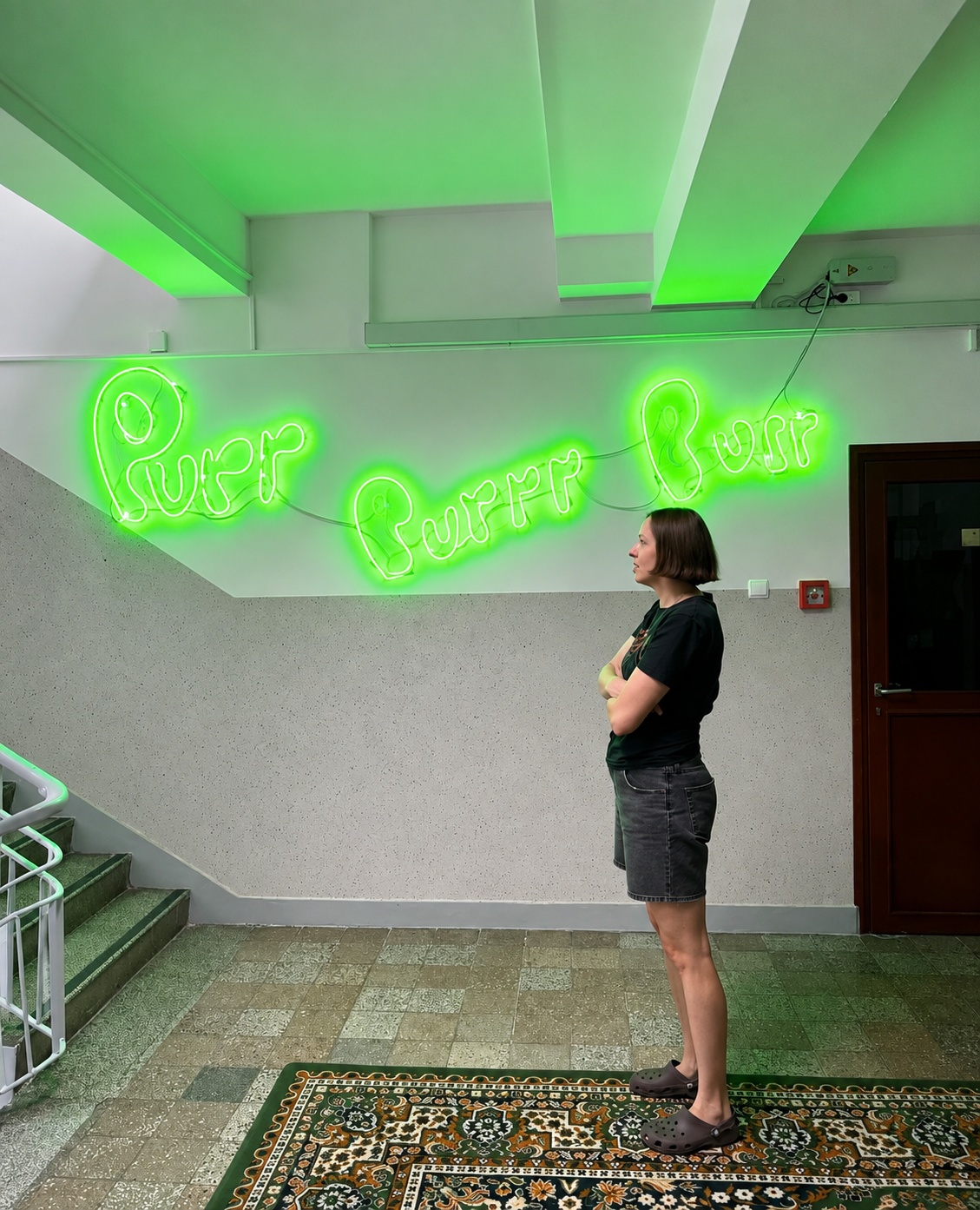 A woman wearing a black T-shirt, black shorts, and black Crocs stands in a hallway with a patterned rug, looking at a neon green sign that says 'Purr Purr Purr' on the wall.