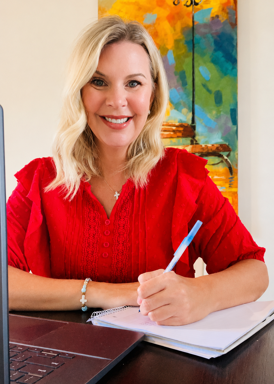 A woman with blonde hair, smiling, wearing a red blouse, sitting at a desk with a laptop, writing in a notebook, with a colorful abstract painting in the background.