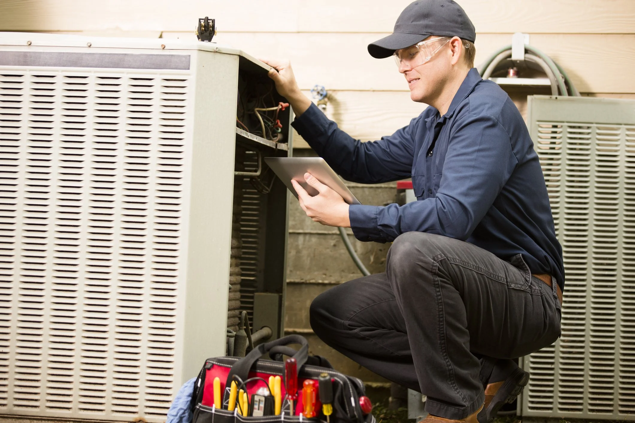 A technician wearing safety glasses and a black cap inspecting an air conditioning unit with a tablet in his hand. A toolbox with tools is on the ground nearby.