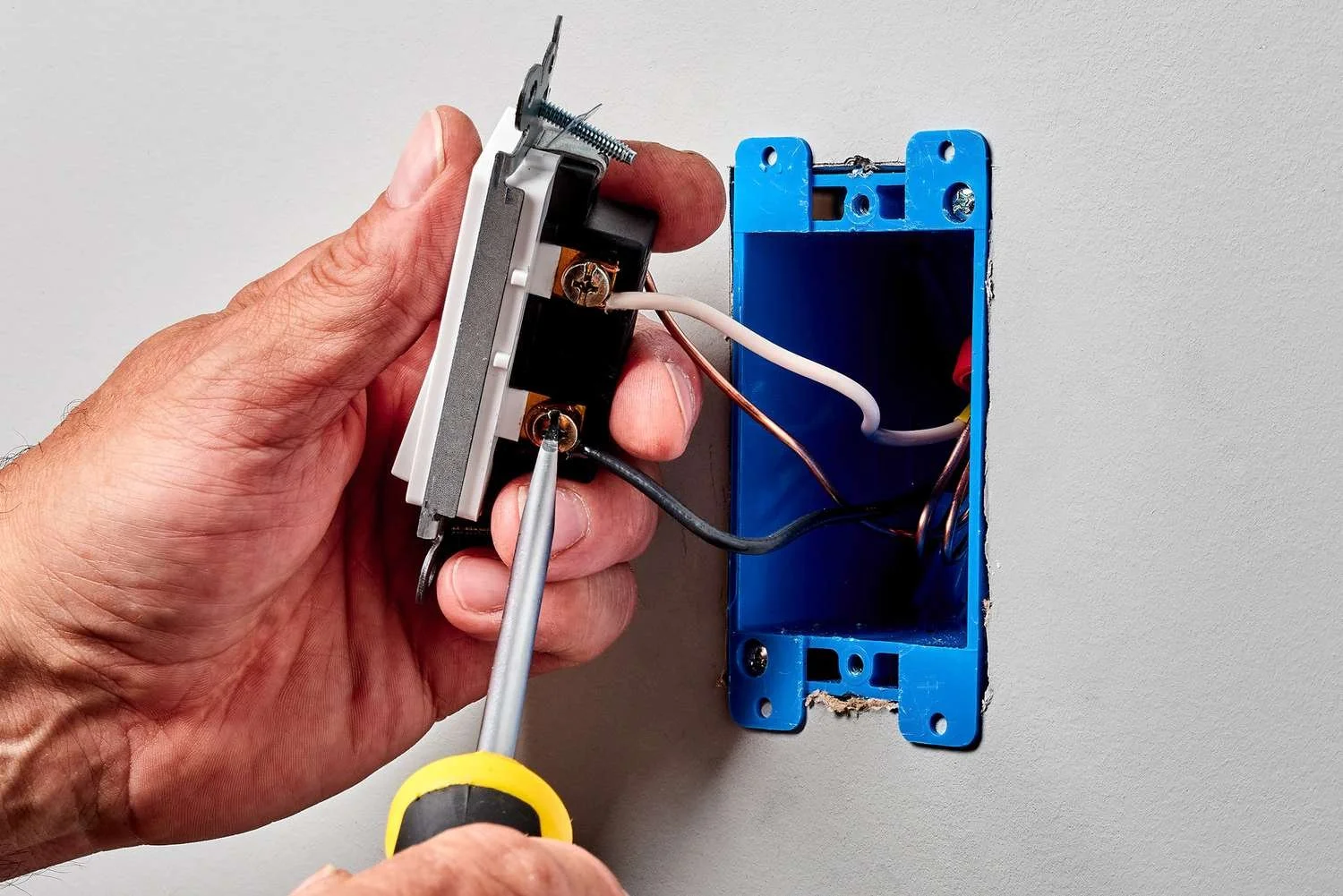 Close-up of a person's hand installing or repairing electrical wiring inside a blue electrical box on a wall.