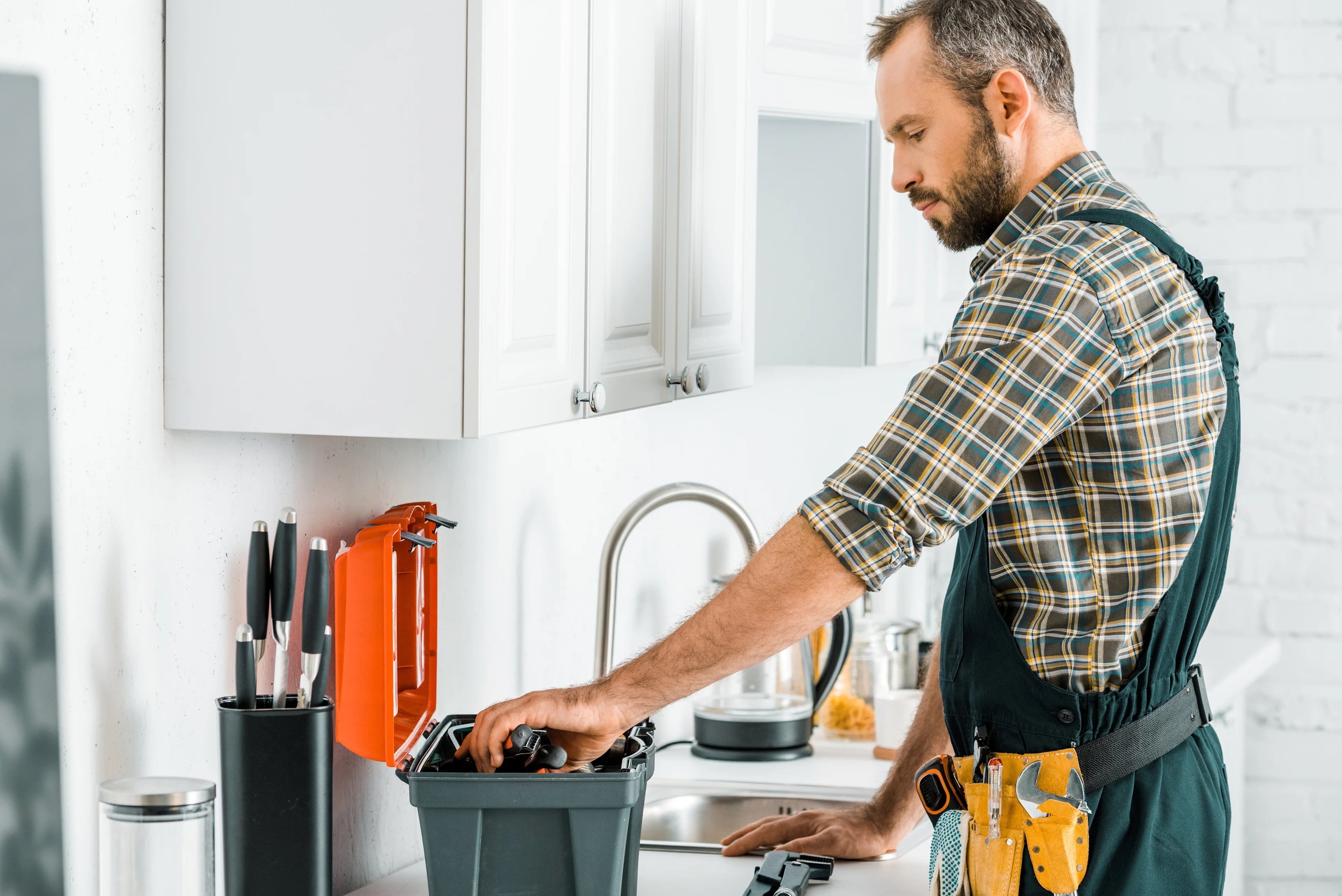 A man wearing a plaid shirt and work apron organizing tools in a kitchen workshop.